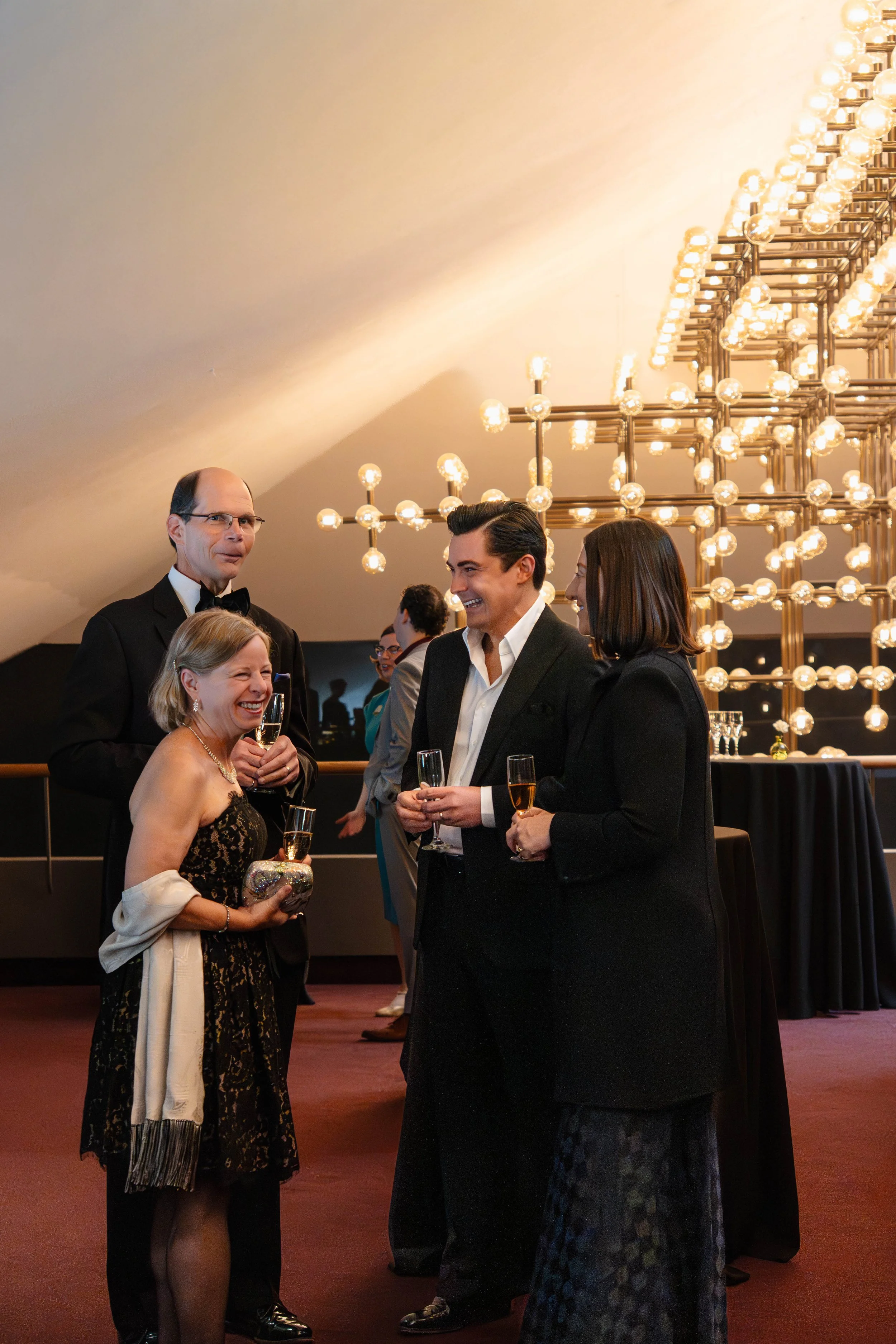 A group of five people dressed in formal attire at a social event, standing and chatting with glasses of champagne in their hands, with a decorative chandelier in the background.