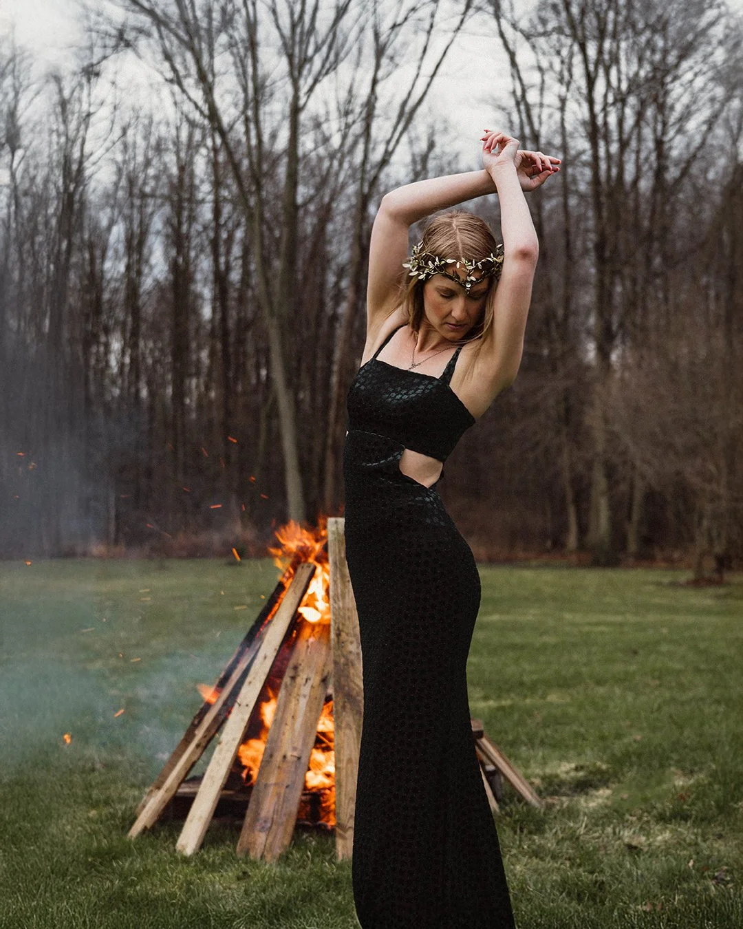 A woman wearing a black dress with cutouts and a floral headband dances outdoors near a bonfire, with leafless trees in the background.