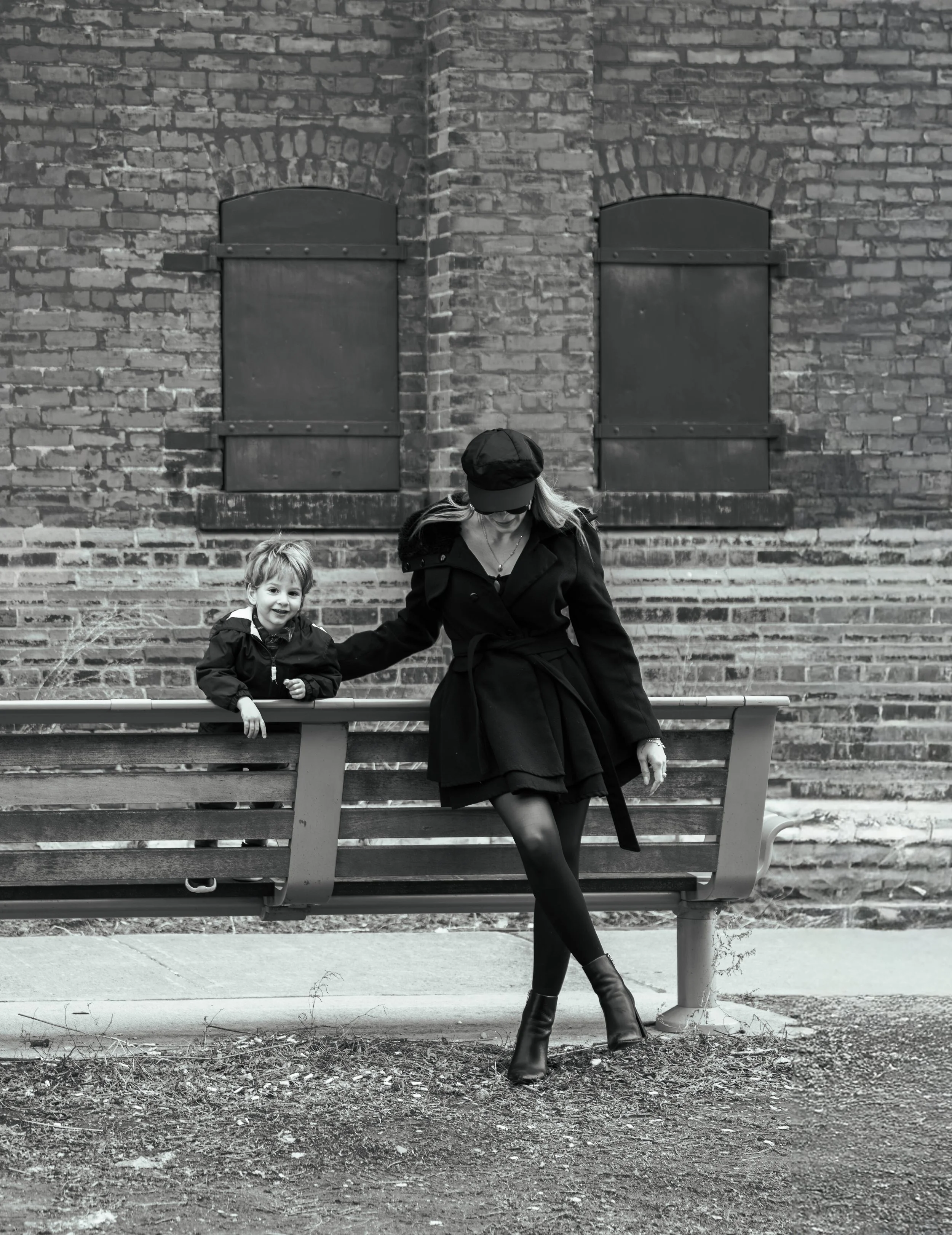Woman and young boy sitting on a park bench in front of a brick wall with two windows. The woman is wearing a black coat, black hat, and black boots, with her head lowered. The boy is smiling, wearing a dark jacket. The ground is dirt with some small