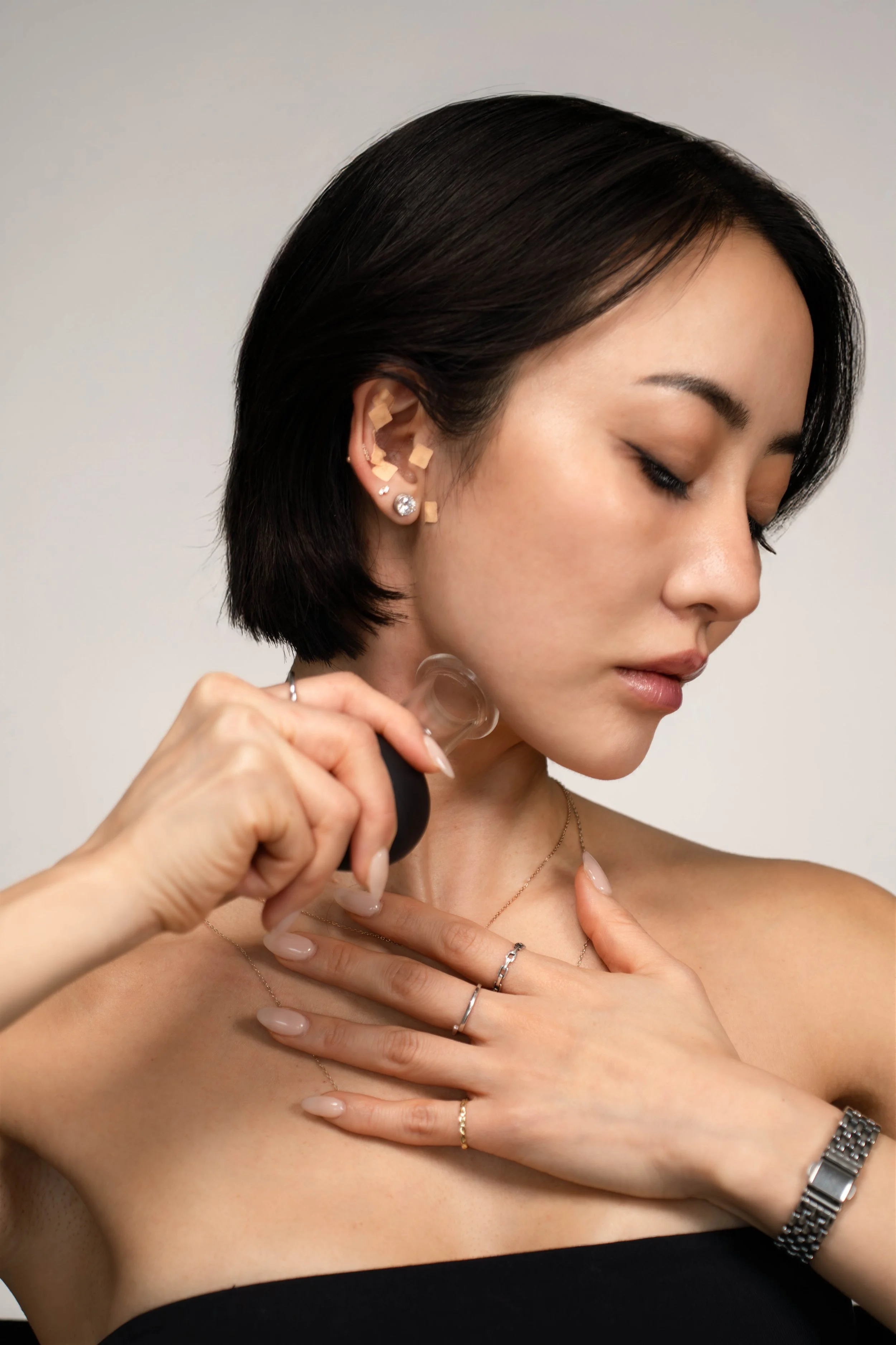 A woman with short black hair is using a skincare device on her neck, wearing jewelry including earrings, rings, a necklace, and a watch, with a neutral background.