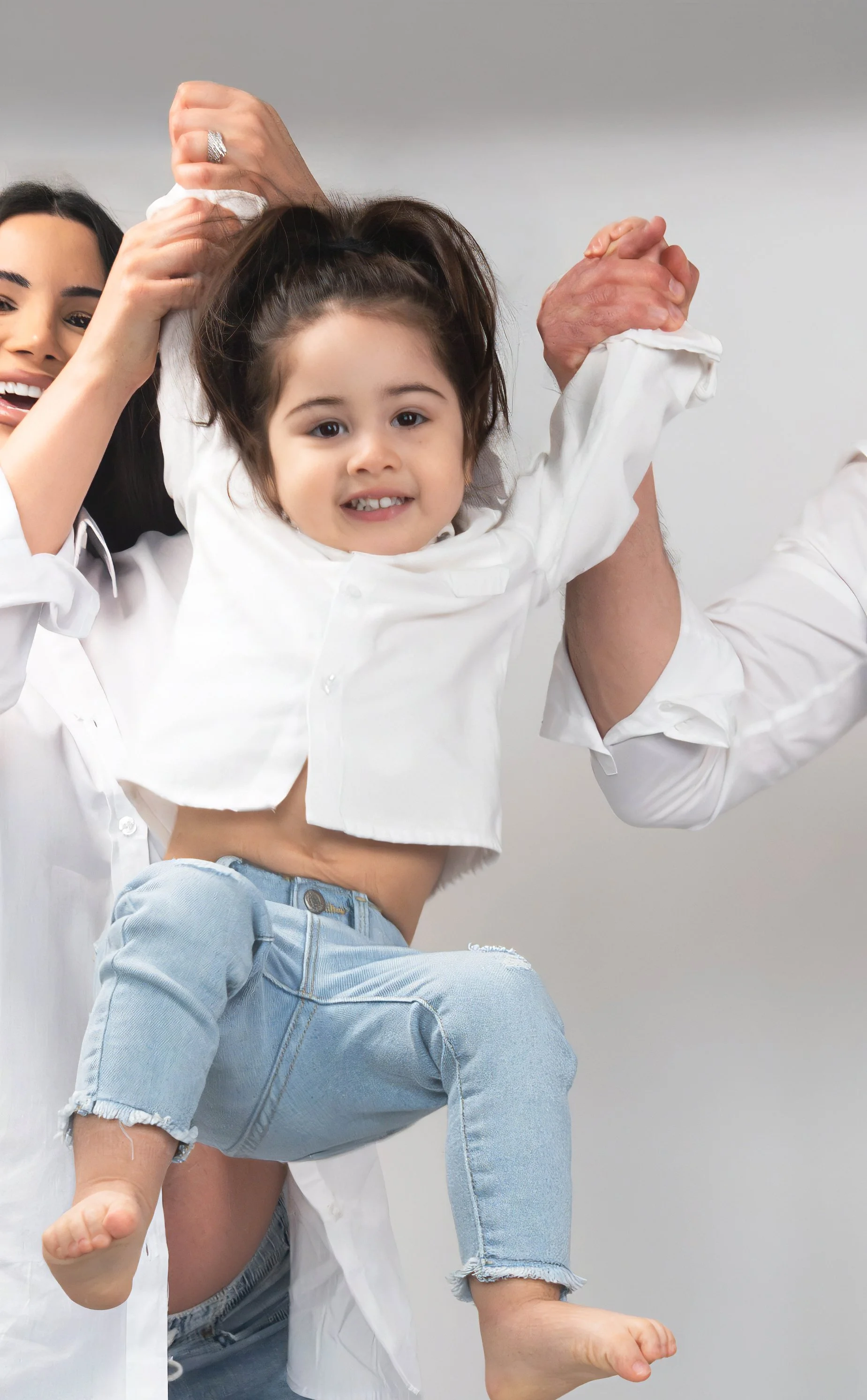 A young girl being lifted by two women, with one woman holding her arm and the other holding her hand. The girl is smiling and wearing a white shirt and ripped jeans. The women are also wearing white shirts, and the background is plain gray.