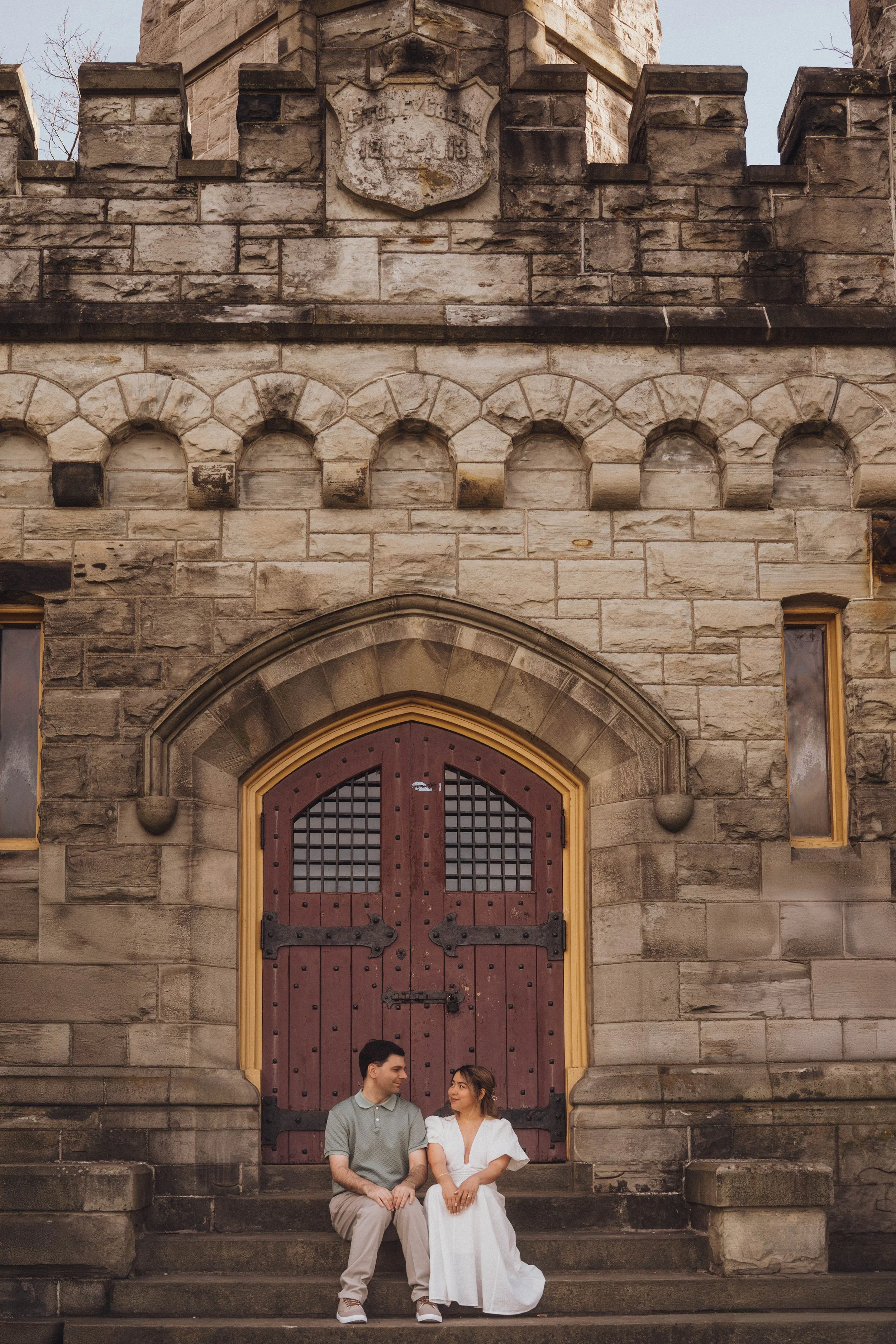 A young man and woman sit on stone stairs in front of a large, historic, Gothic-style stone building with a arched wooden door and decorative stonework, engaging in conversation. The man is wearing a light gray polo shirt and beige pants, while the w