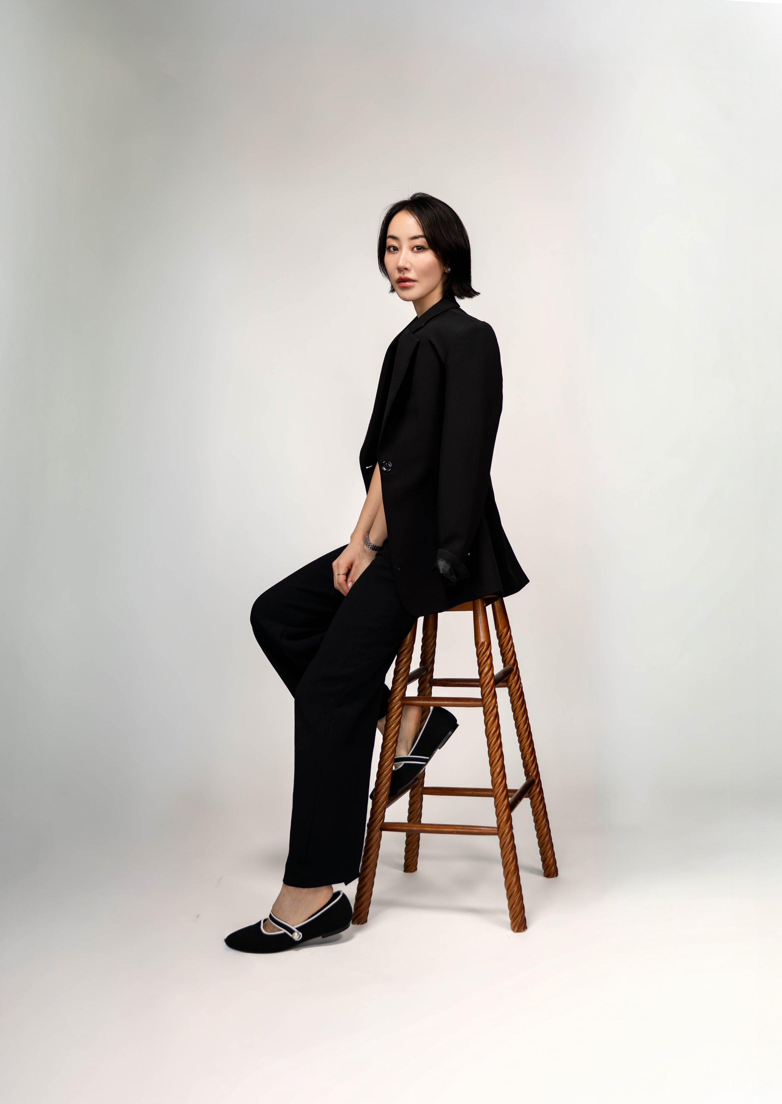 A woman dressed in black, sitting sideways on a wooden barstool, against a plain white background.