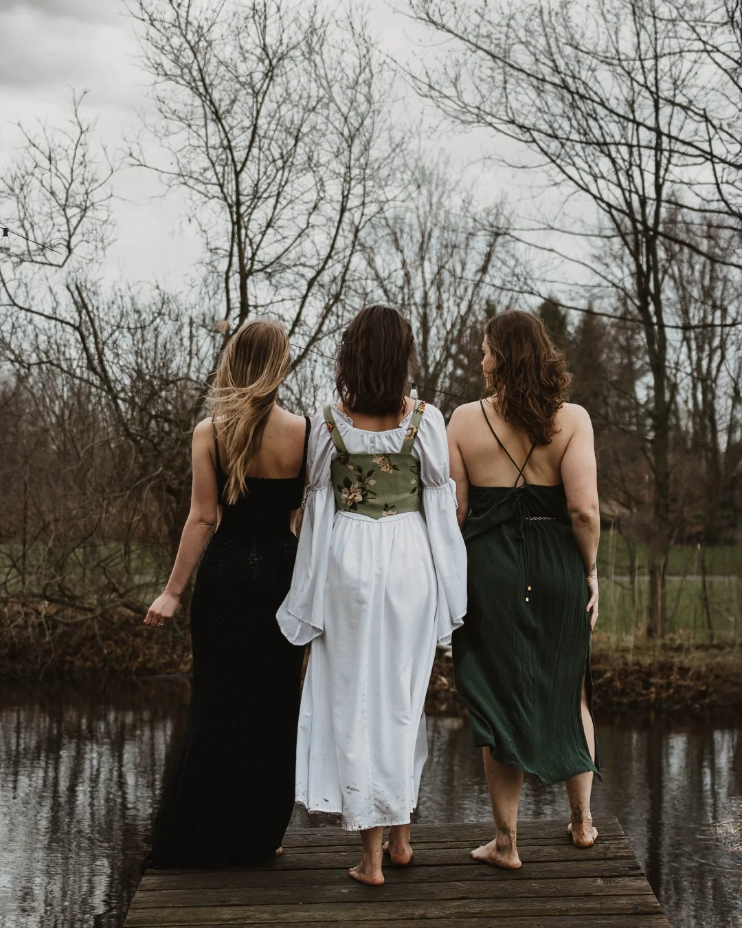 Three women standing on a wooden dock with their backs to the camera, overlooking a small pond with bare trees in the background, during overcast weather.