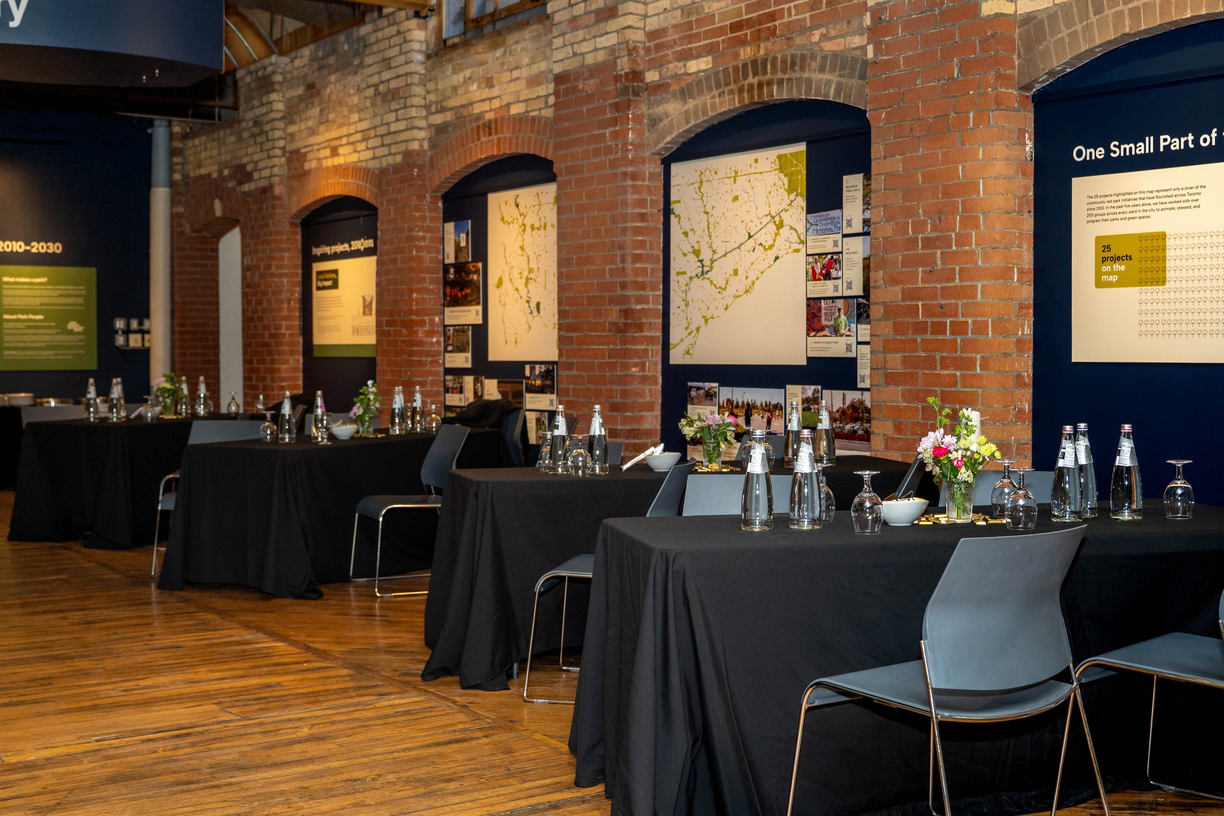 Event space with tables covered in black tablecloths, set with water bottles, glasses, bowls, and floral centerpieces, against a brick wall with informational posters.