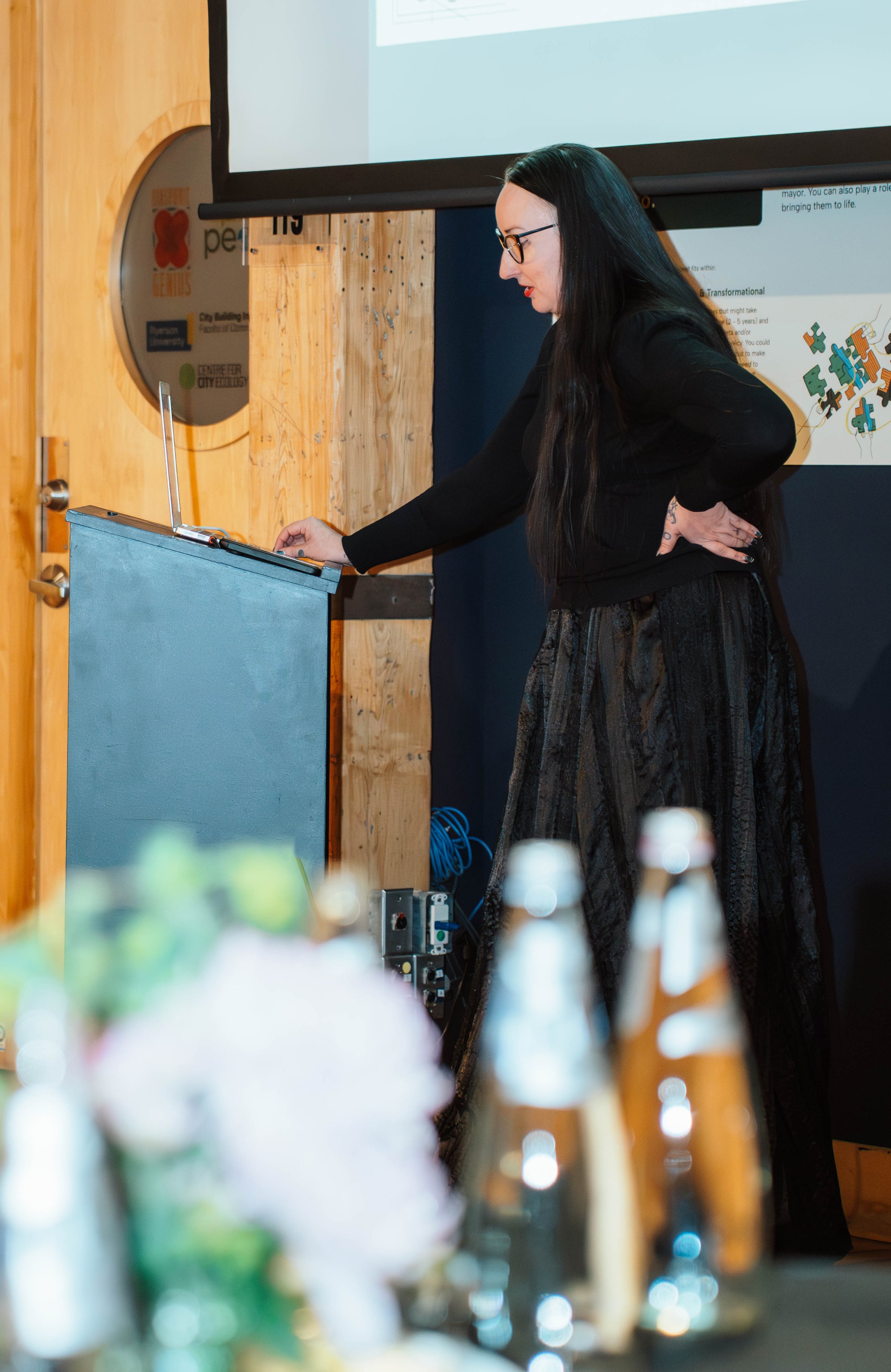 A woman with long black hair and glasses, dressed in black, is standing at a podium, looking at a laptop, during a presentation in a conference room.