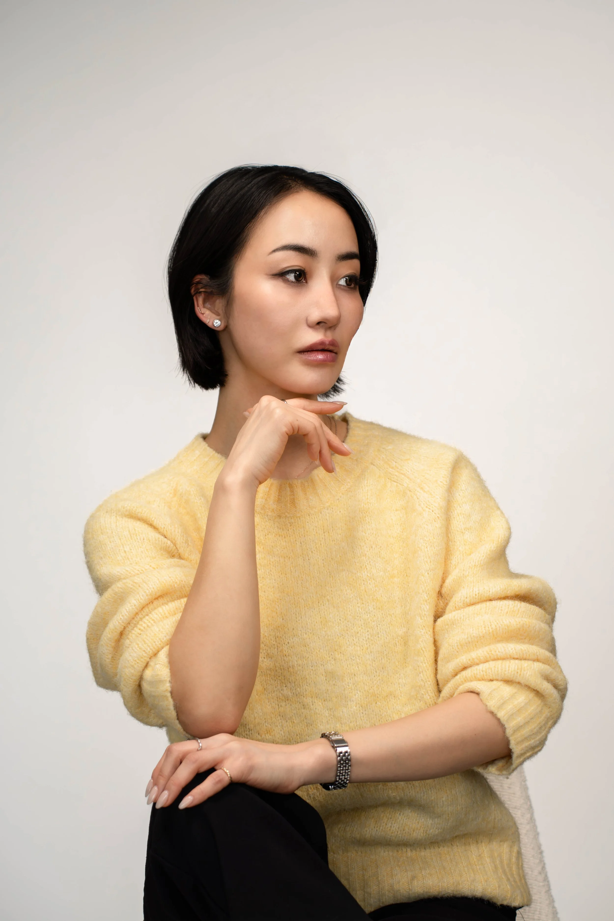 A woman with short black hair, wearing a yellow sweater, looking to the side with her hand near her chin, against a plain light background.