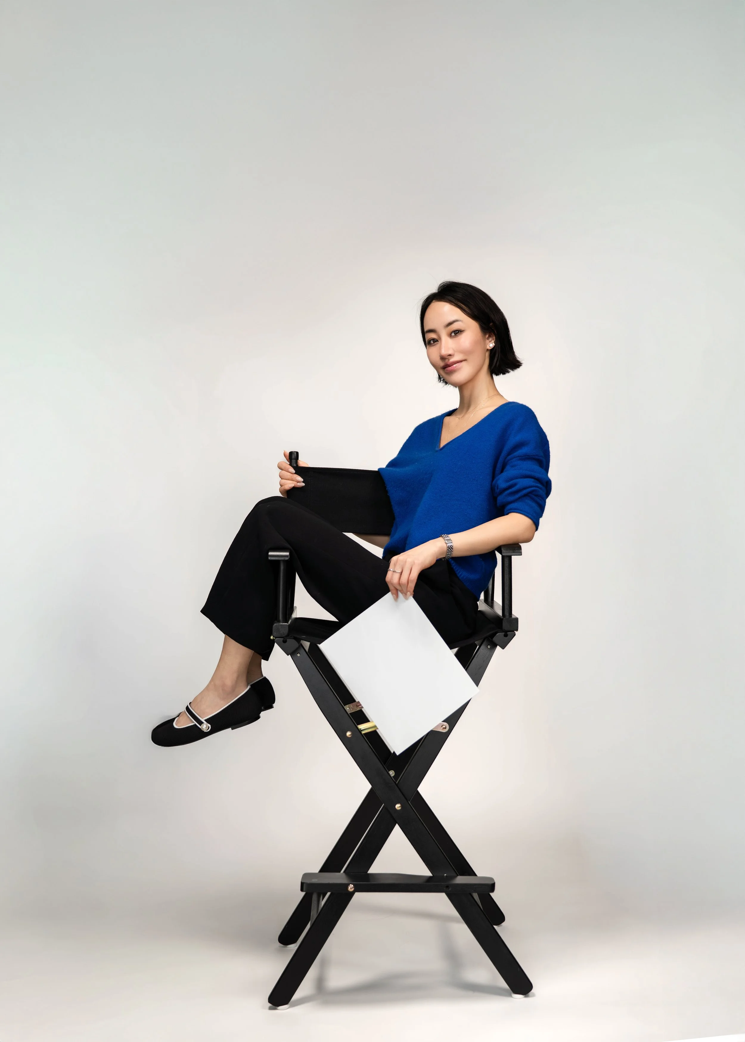 A woman with black hair, wearing a blue sweater and black pants, sitting on a director's chair against a plain white background, holding a black folder, with a white sheet of paper hanging from her leg.