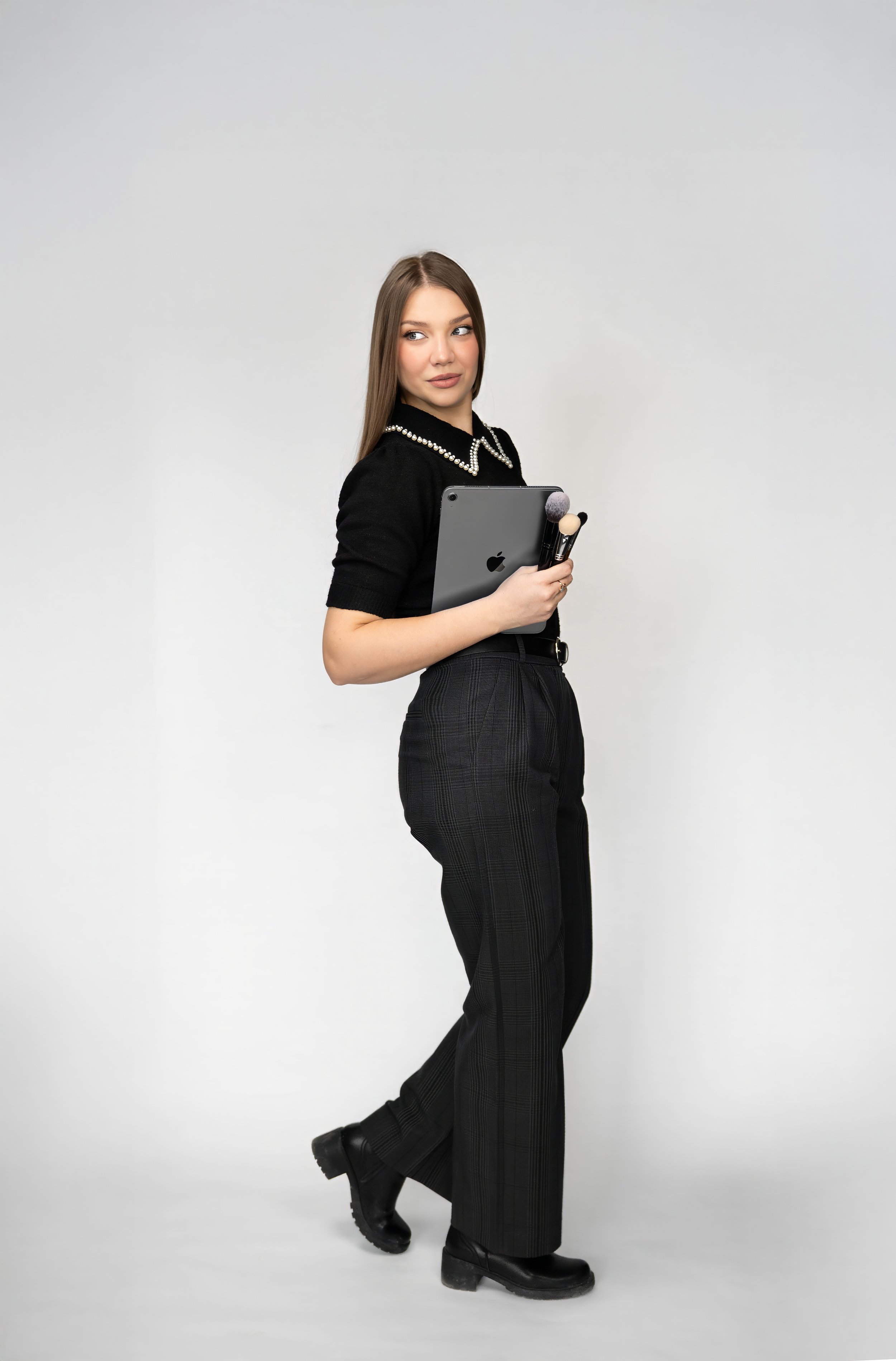 A young woman holding an iPad and makeup brushes, wearing a black top with pearl embellishments and black pants, standing against a plain white background.