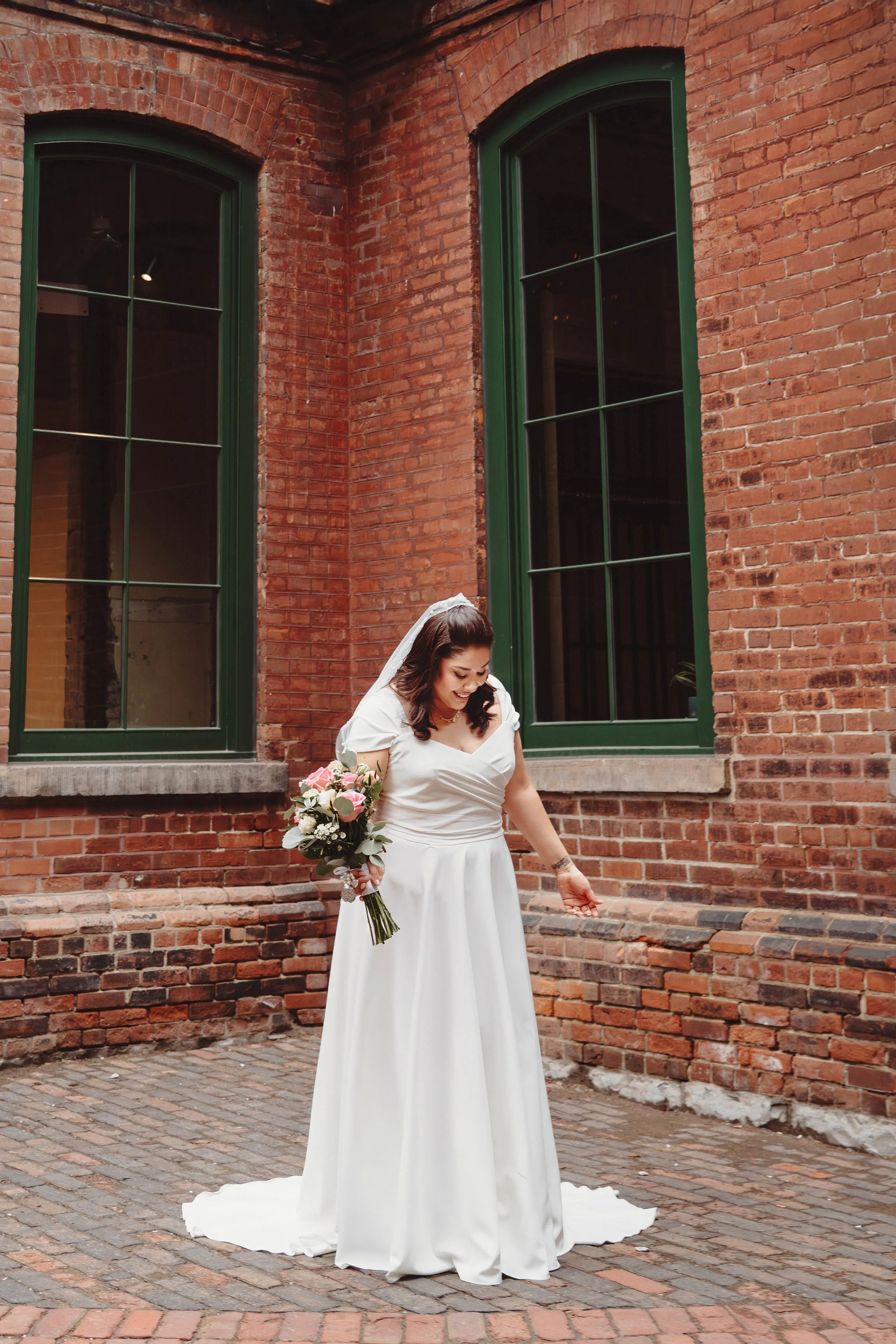 Bridal bride in white wedding dress holding bouquet of pink and white roses, standing outside against a brick wall with large green-framed windows.