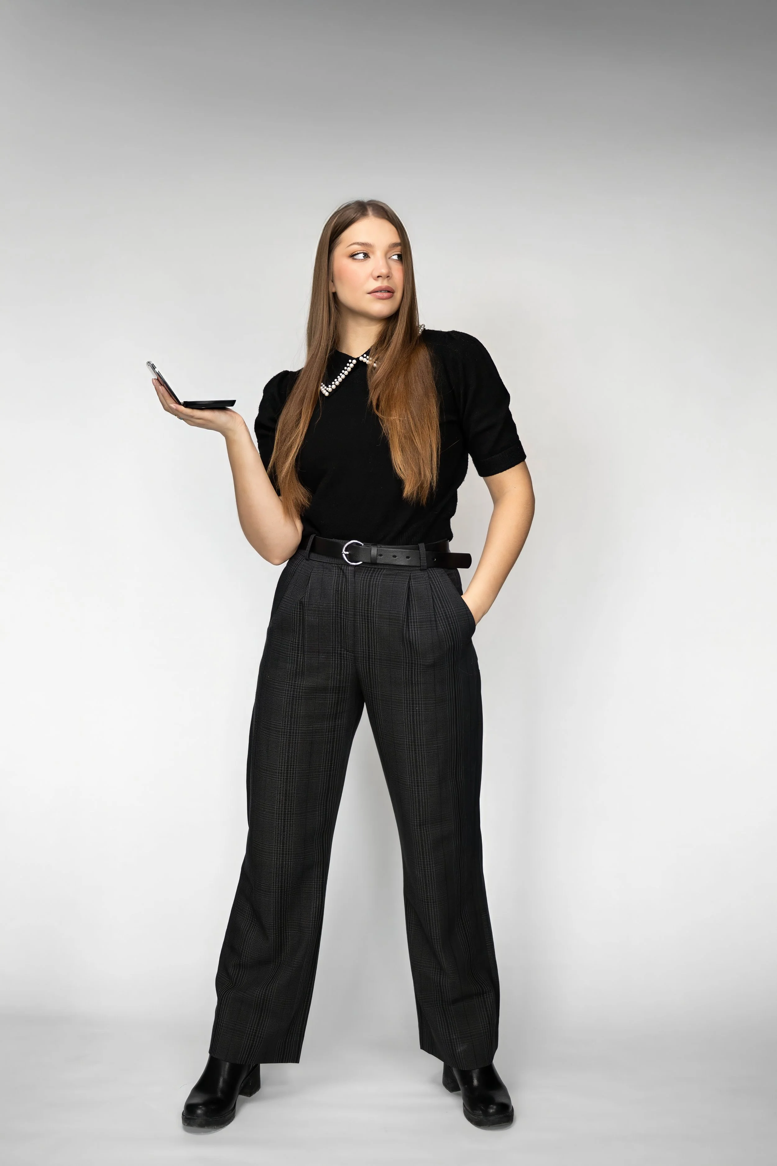A young woman with long brown hair stands against a plain white background. She is dressed in a black top with a pearl necklace, plaid gray trousers, and black boots. She holds a smartphone in her right hand and looks to her left.