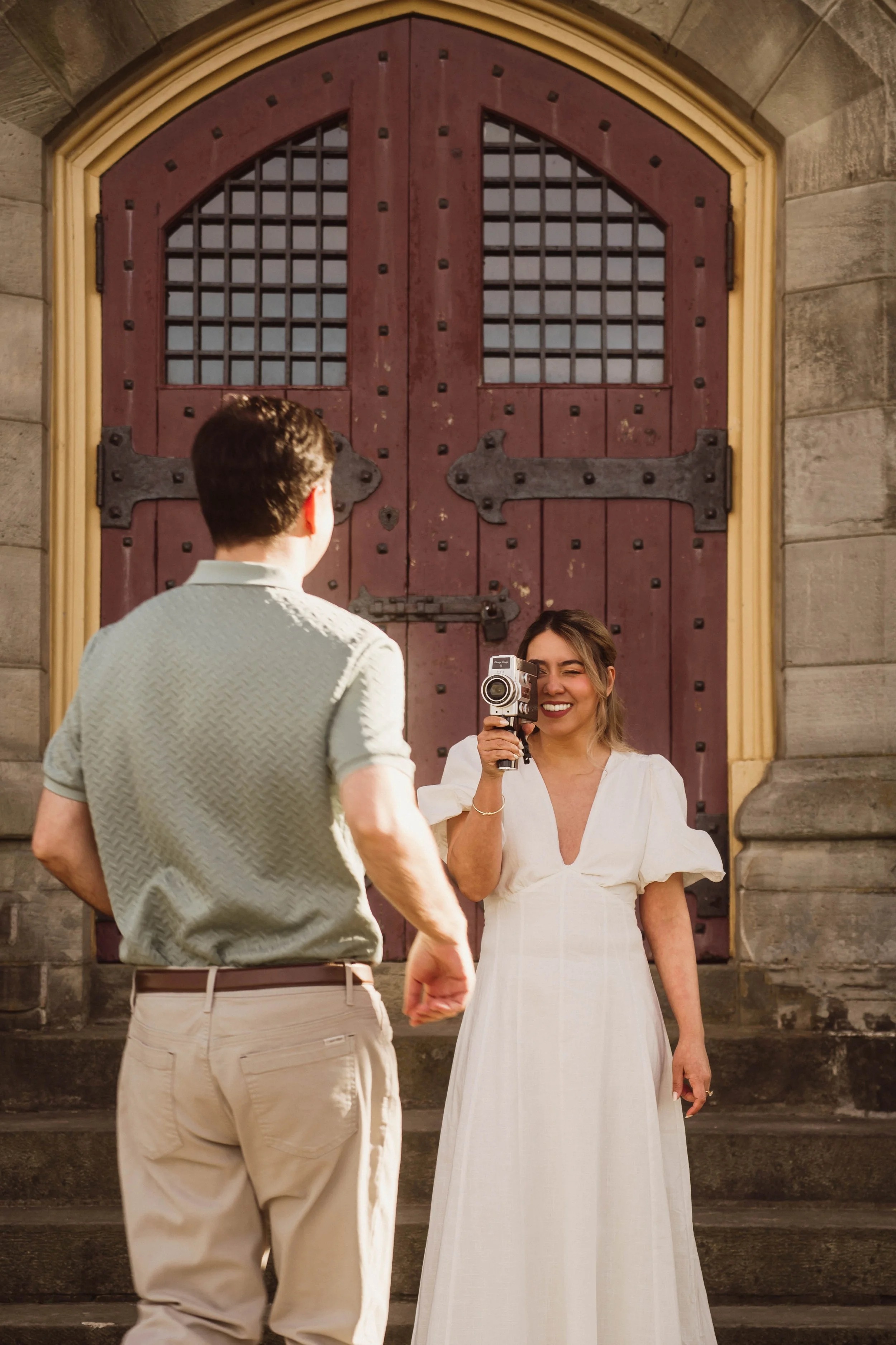 Bride-to-be smiling and filming her partner with a vintage camera during an engagement session at Battlefield Park in Hamilton, Ontario, in front of historic stone architecture