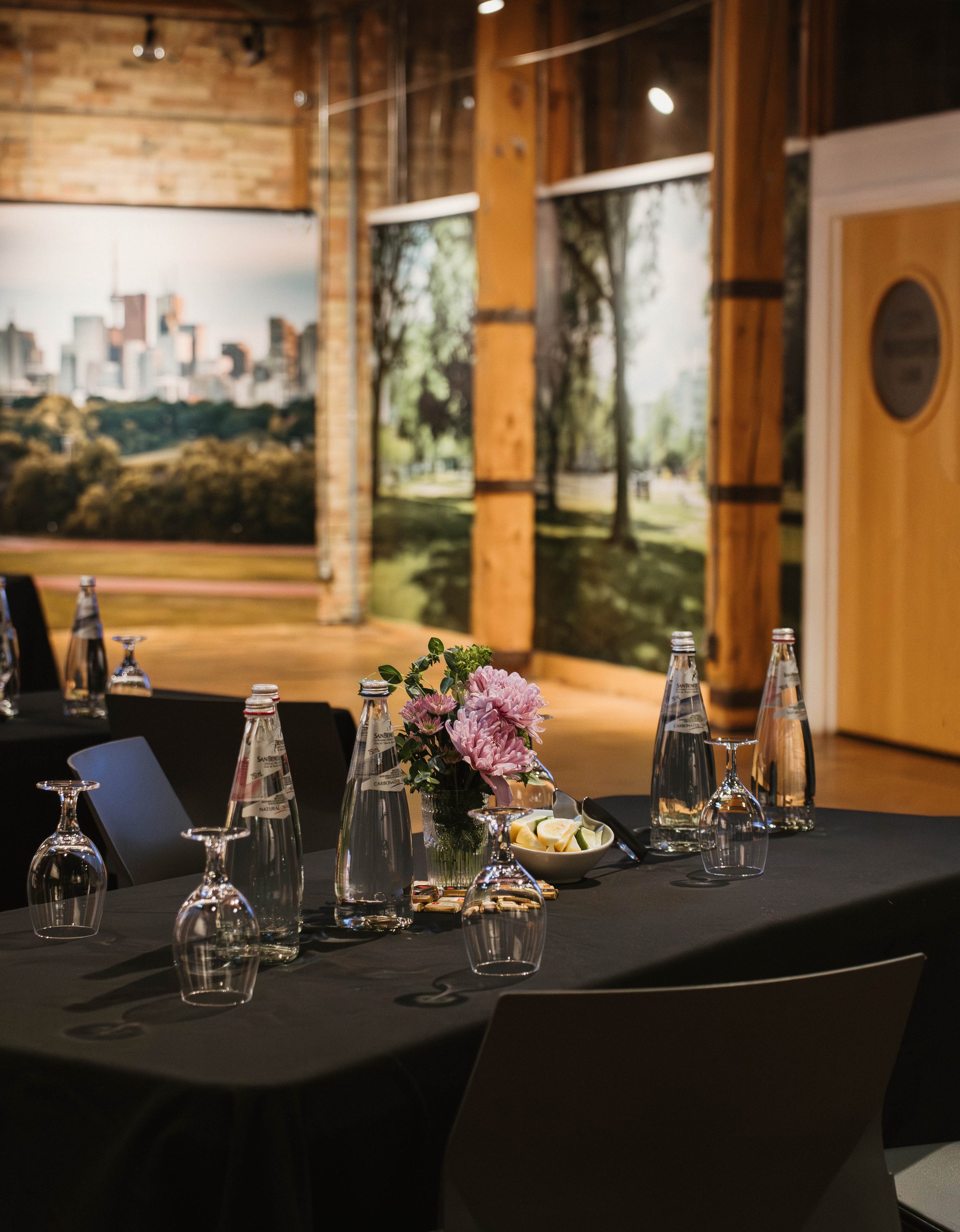 A decorated table set for a meal with wine glasses, water bottles, a pink flower centerpiece, and lemon slices in a restaurant with wood and brick interior and large wall photos of a park and cityscape.
