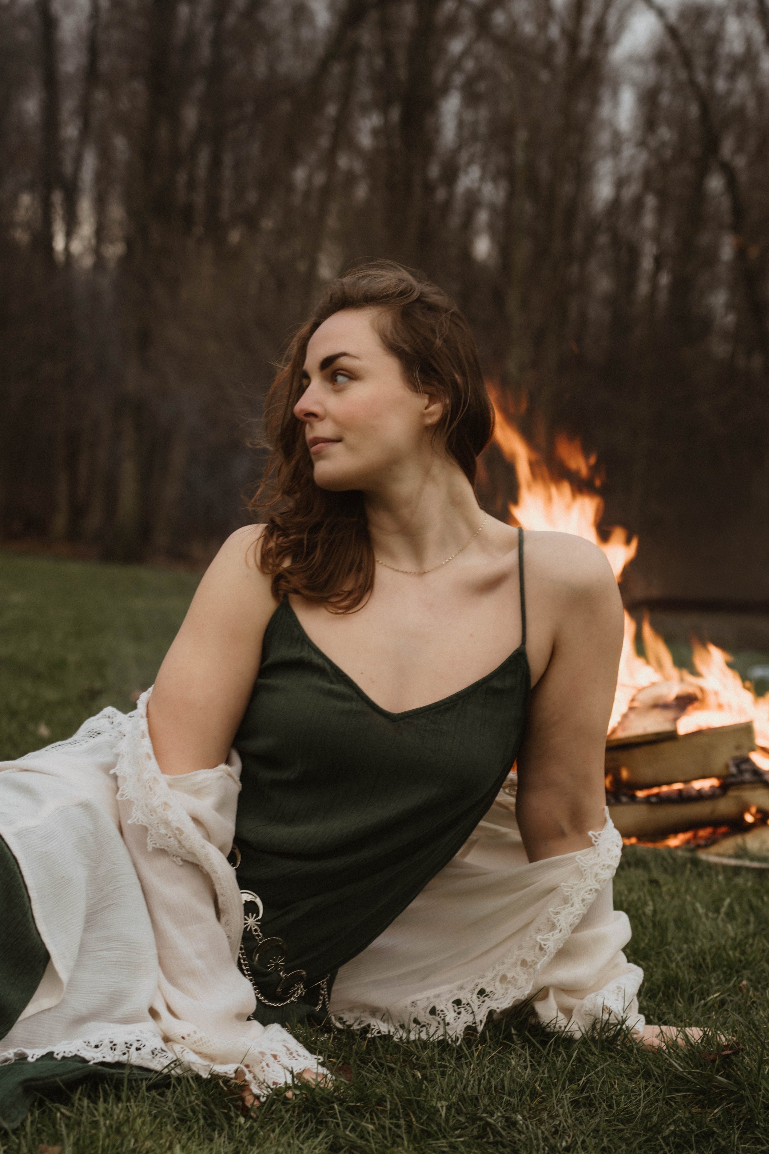 A young woman with wavy auburn hair lying on grass by a campfire in a wooded area, wearing a dark green top and a cream-colored jacket with lace details.