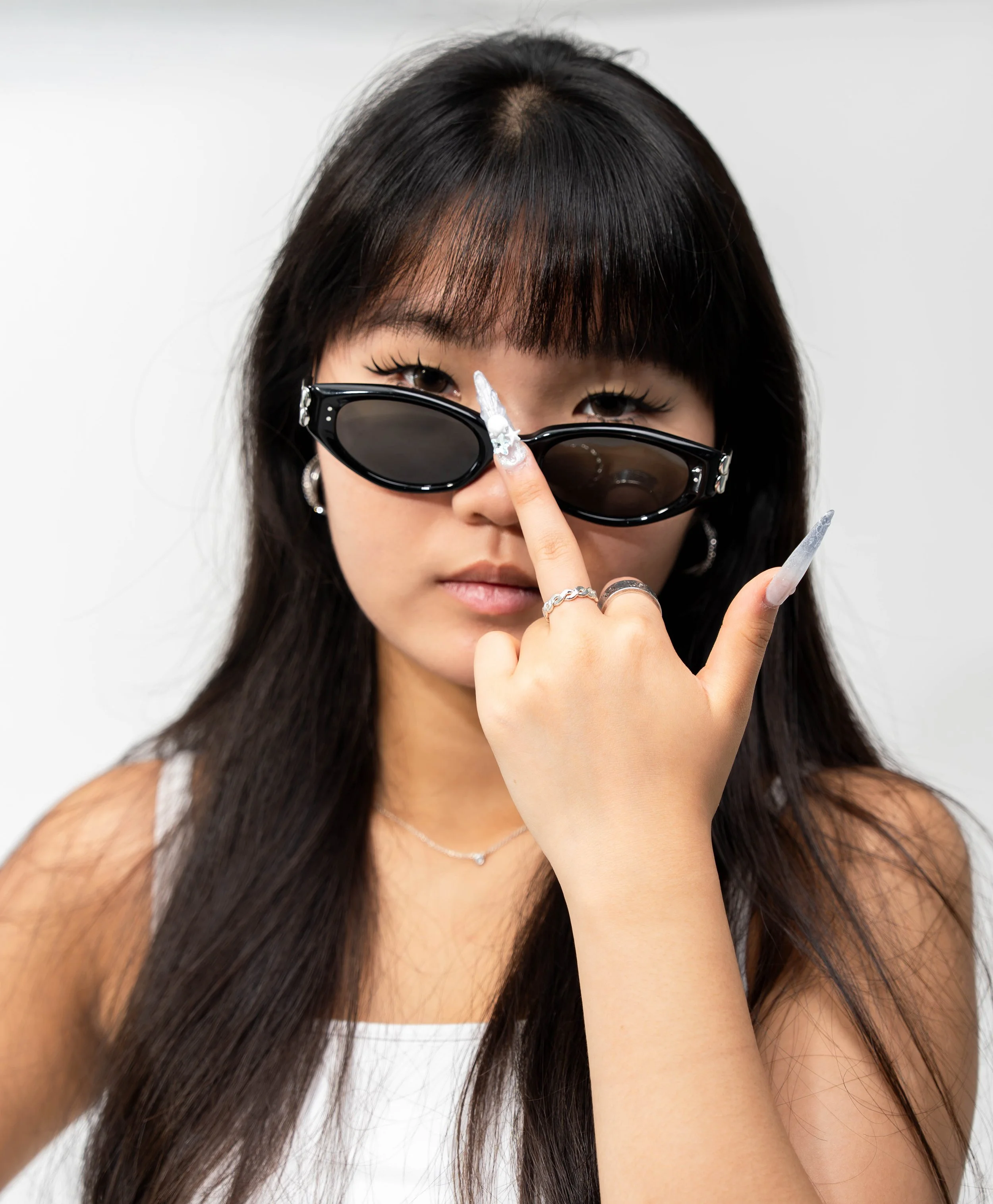 A woman with long dark hair and bangs wearing black sunglasses, holding a cigarette, with a white background.