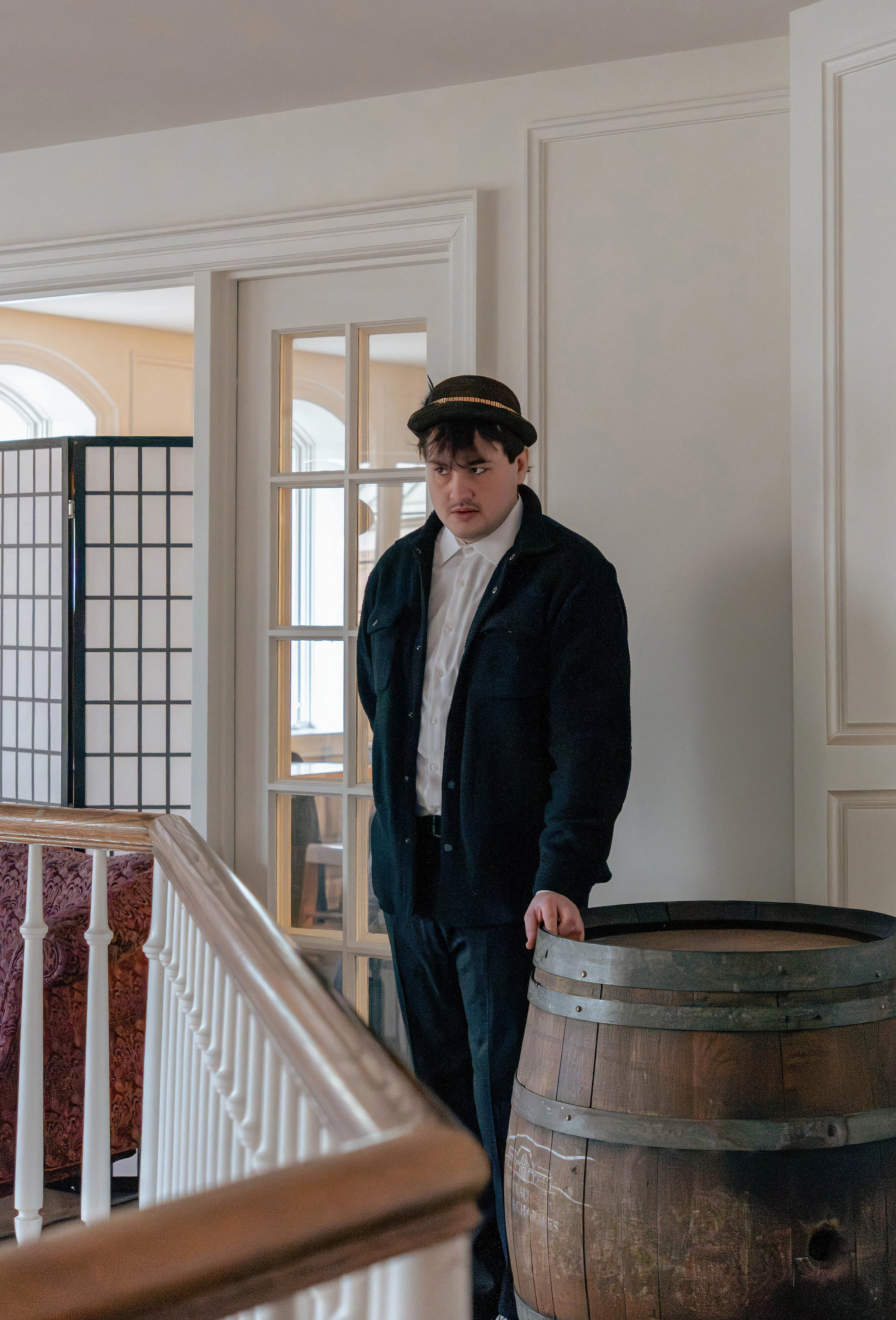 A young man standing indoors next to a large wooden barrel, wearing a dark jacket, white shirt, and a hat, with a serious expression.