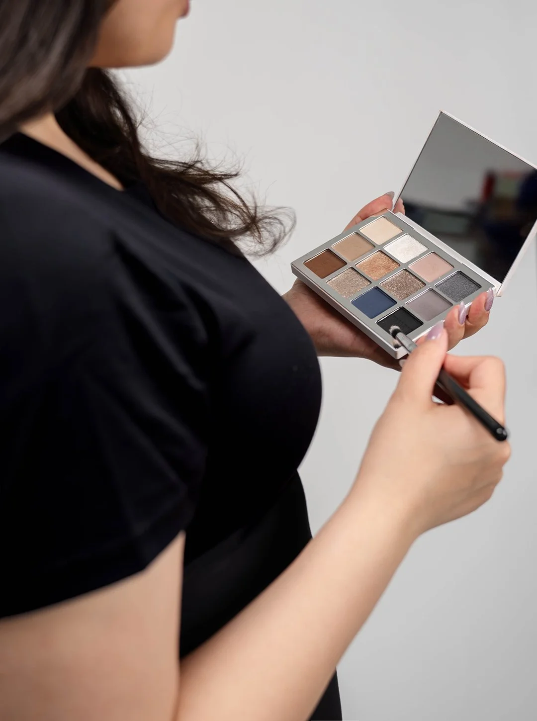A woman holding an open eyeshadow palette with multiple colors and a mirror, applying makeup with a brush.