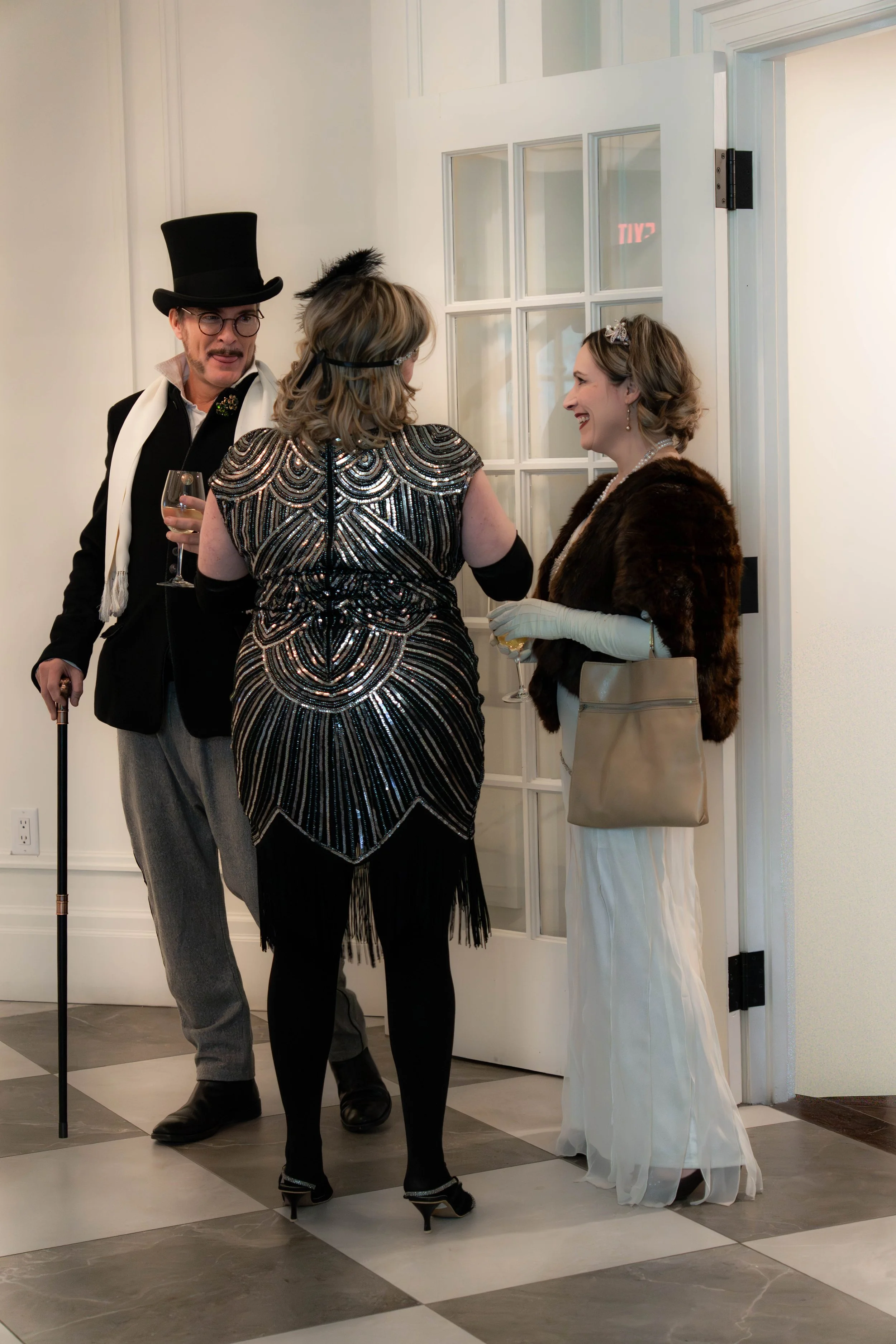 Three people dressed in vintage or costume attire, engaged in conversation at an indoor social event. Two women are holding drinks, and a man with a cane is standing nearby.