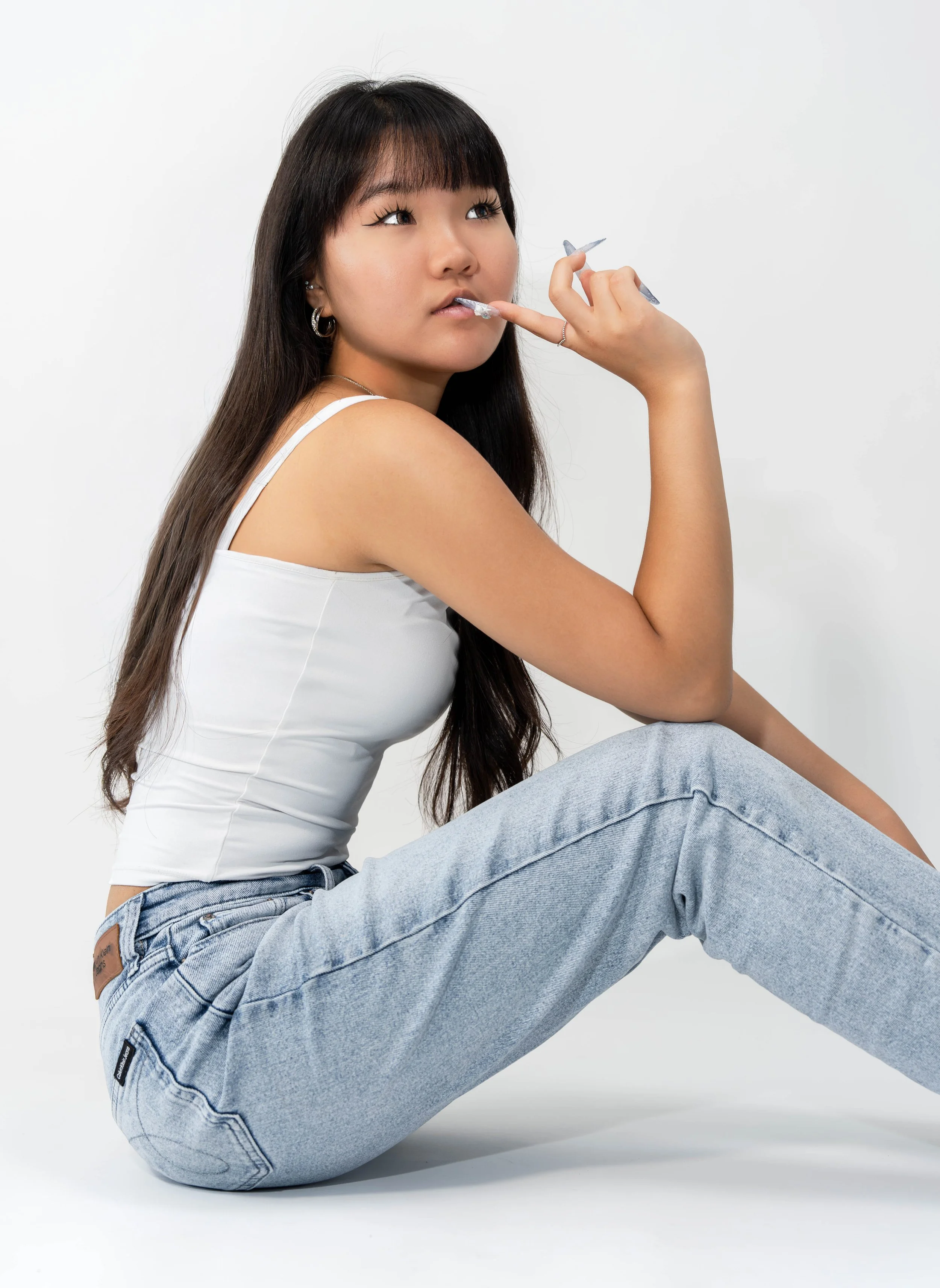 Young woman with long dark hair and bangs, wearing a white tank top and light blue jeans, sitting on the floor against a plain white background, looking upwards with her finger near her lips, in a relaxed pose.