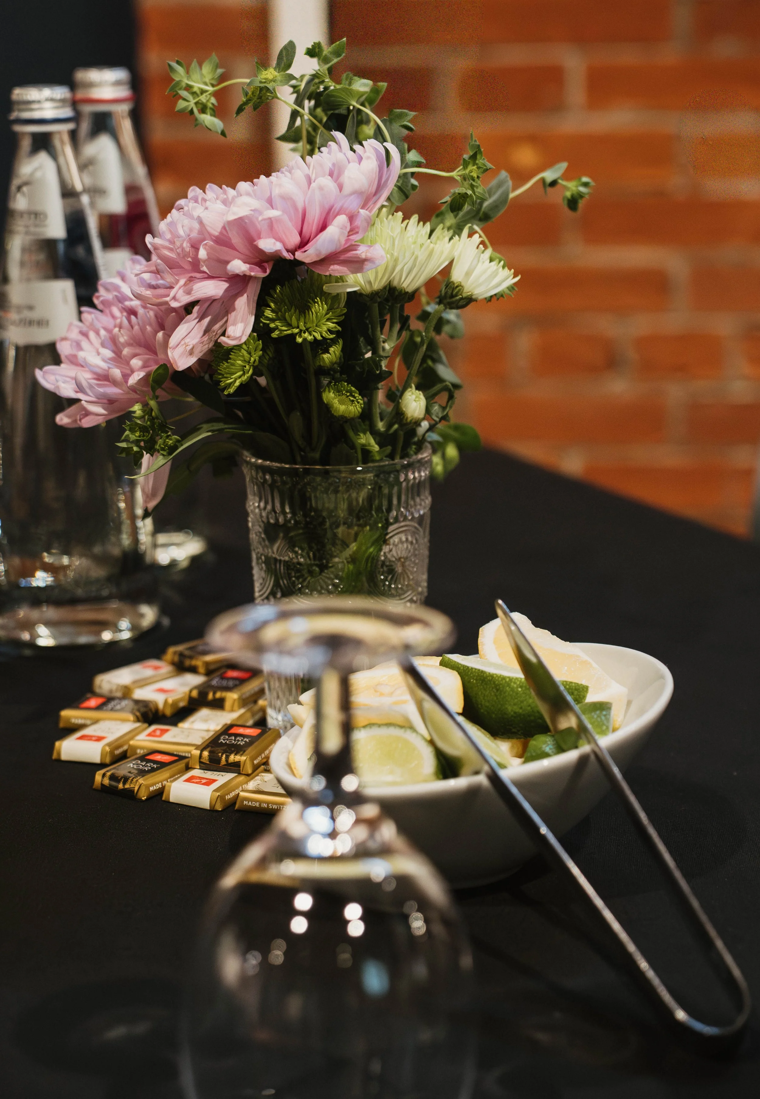 A floral arrangement with pink, white, and green flowers in a glass vase, placed on a table with small chocolate bars, a bowl of lime wedges, and tongs. A blurred wine glass and bottles are in the foreground, and a brick wall is in the background.