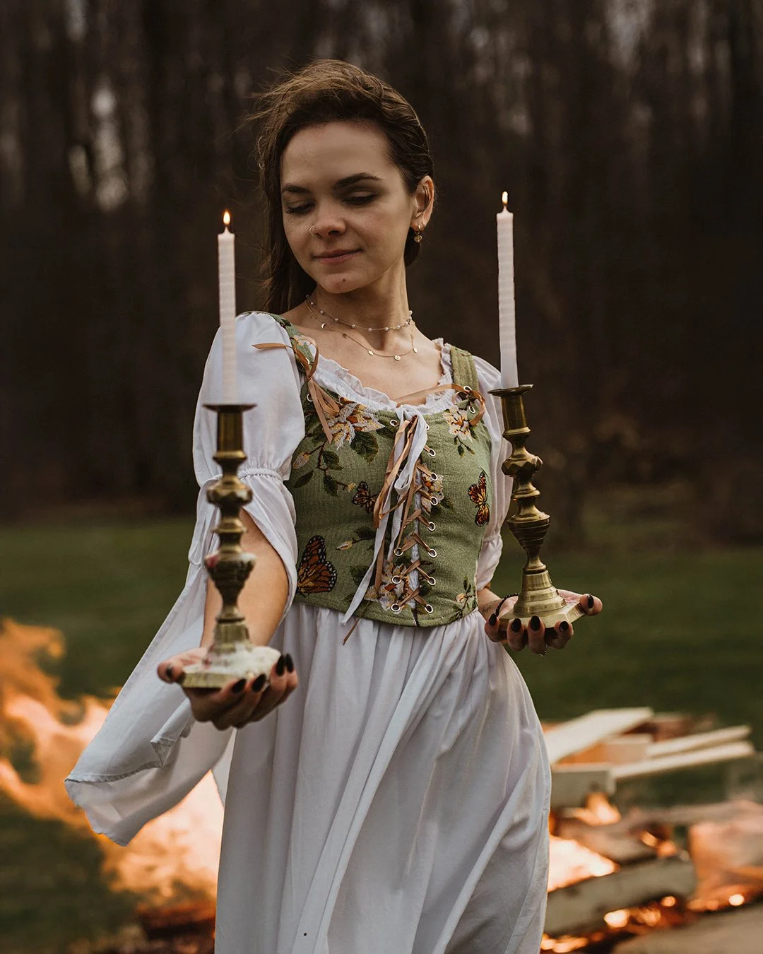 Young woman in a white dress and floral embroidered vest holding two lit candlesticks outdoors near a fire.