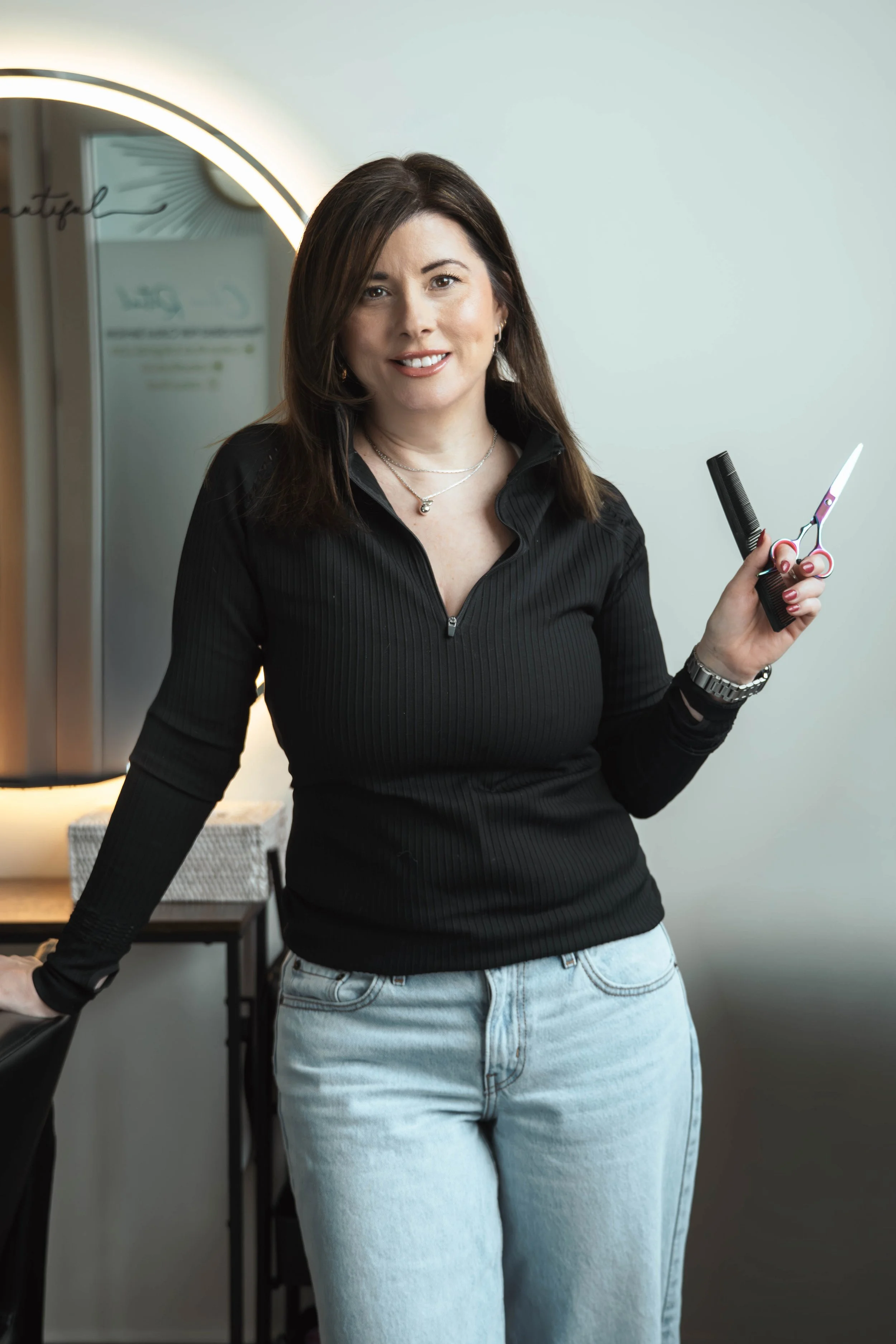 A woman with brown hair smiling, holding scissors and a comb, in a salon or beauty studio.
