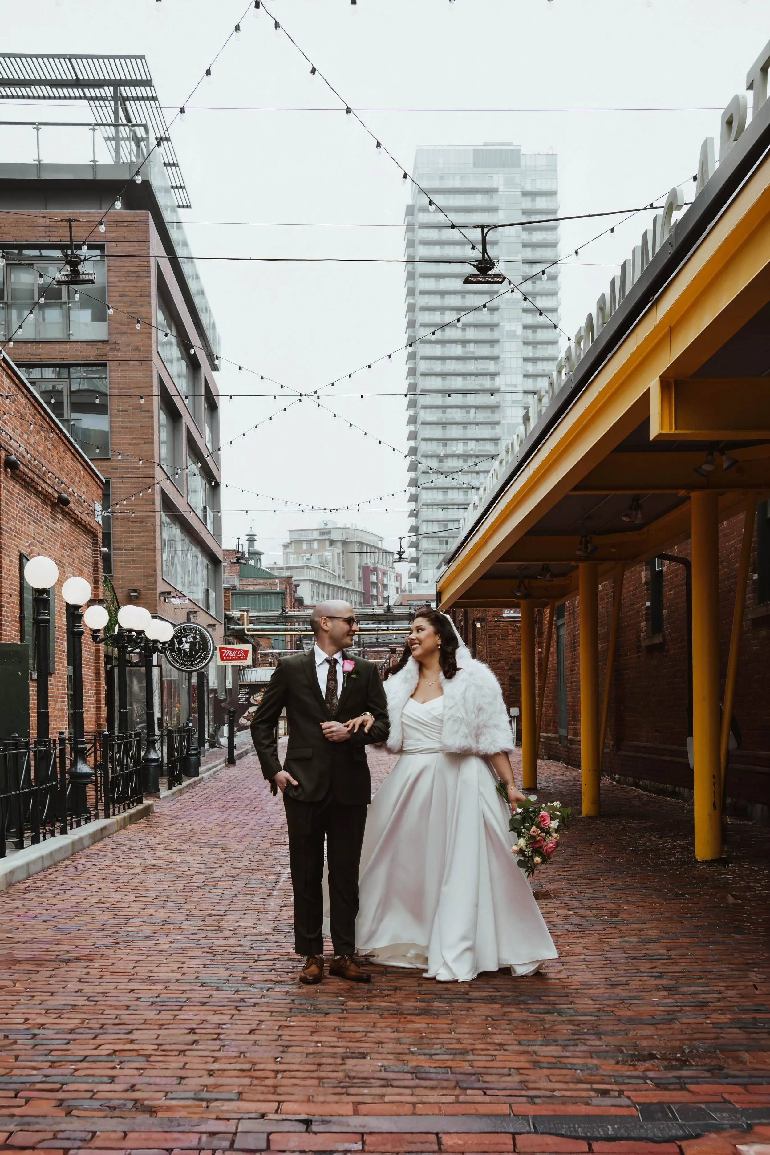 A bride and groom walking arm in arm on a brick sidewalk, smiling at each other in an urban setting with buildings, string lights, and outdoor fixtures.
