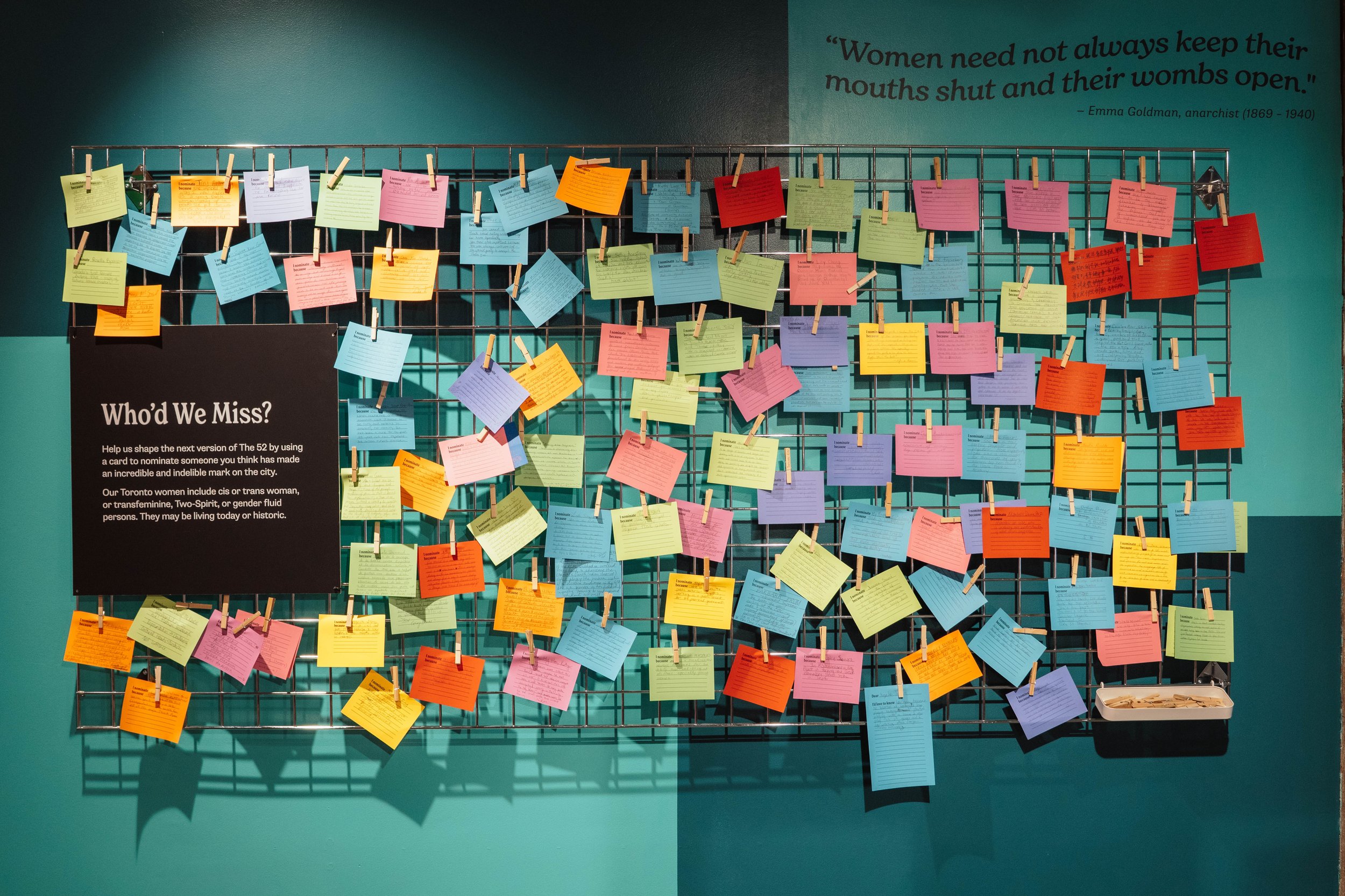 Colorful notes attached to a black wire grid with clothespins, part of an interactive display titled 'Who'd We Miss?' on a teal wall, encouraging visitors to nominate influential or historic women, with a quote from Emma Goldman.