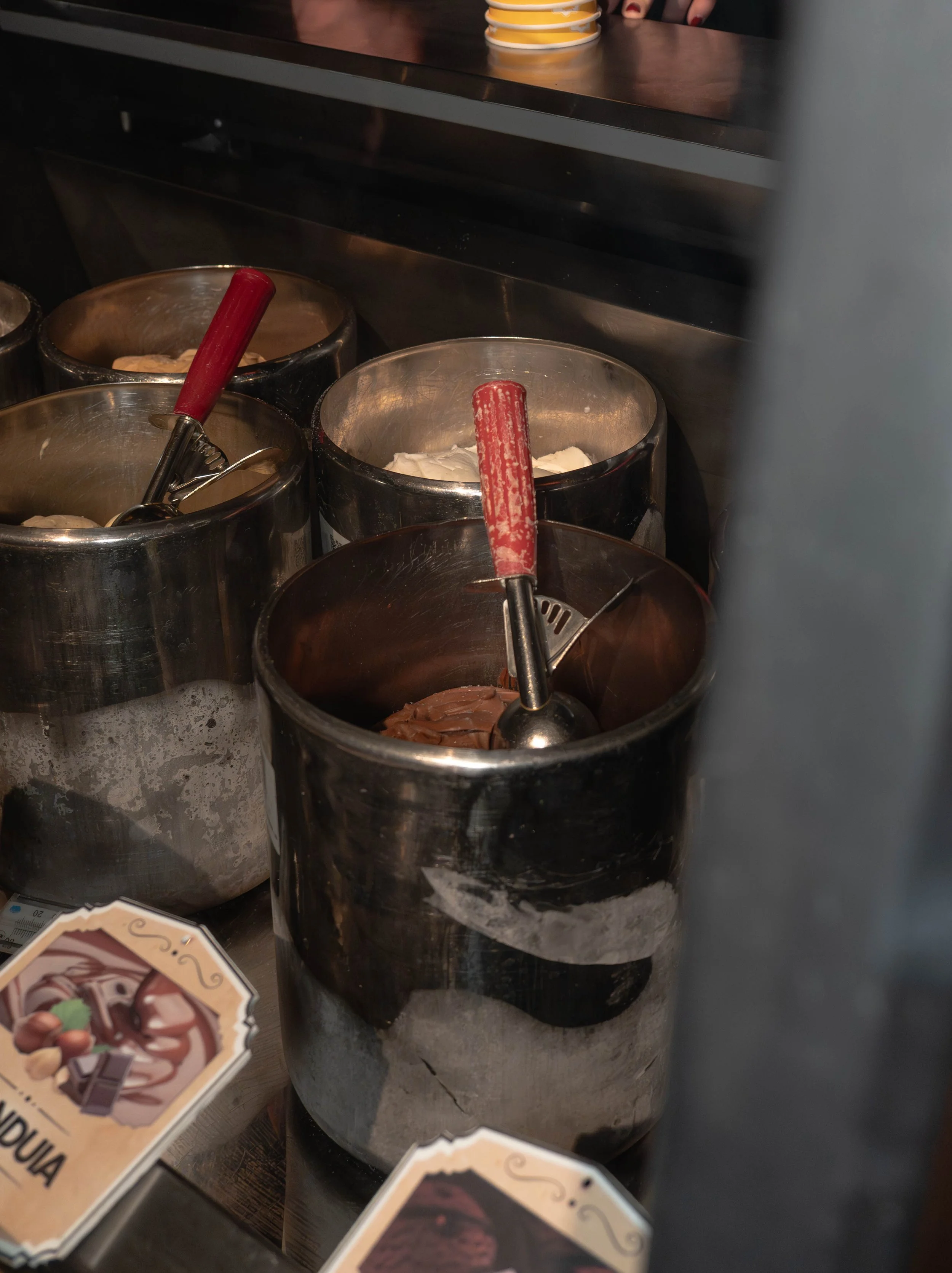 Containers of various toppings and chocolates, with a scoop in one of the containers, likely at an ice cream or dessert shop.