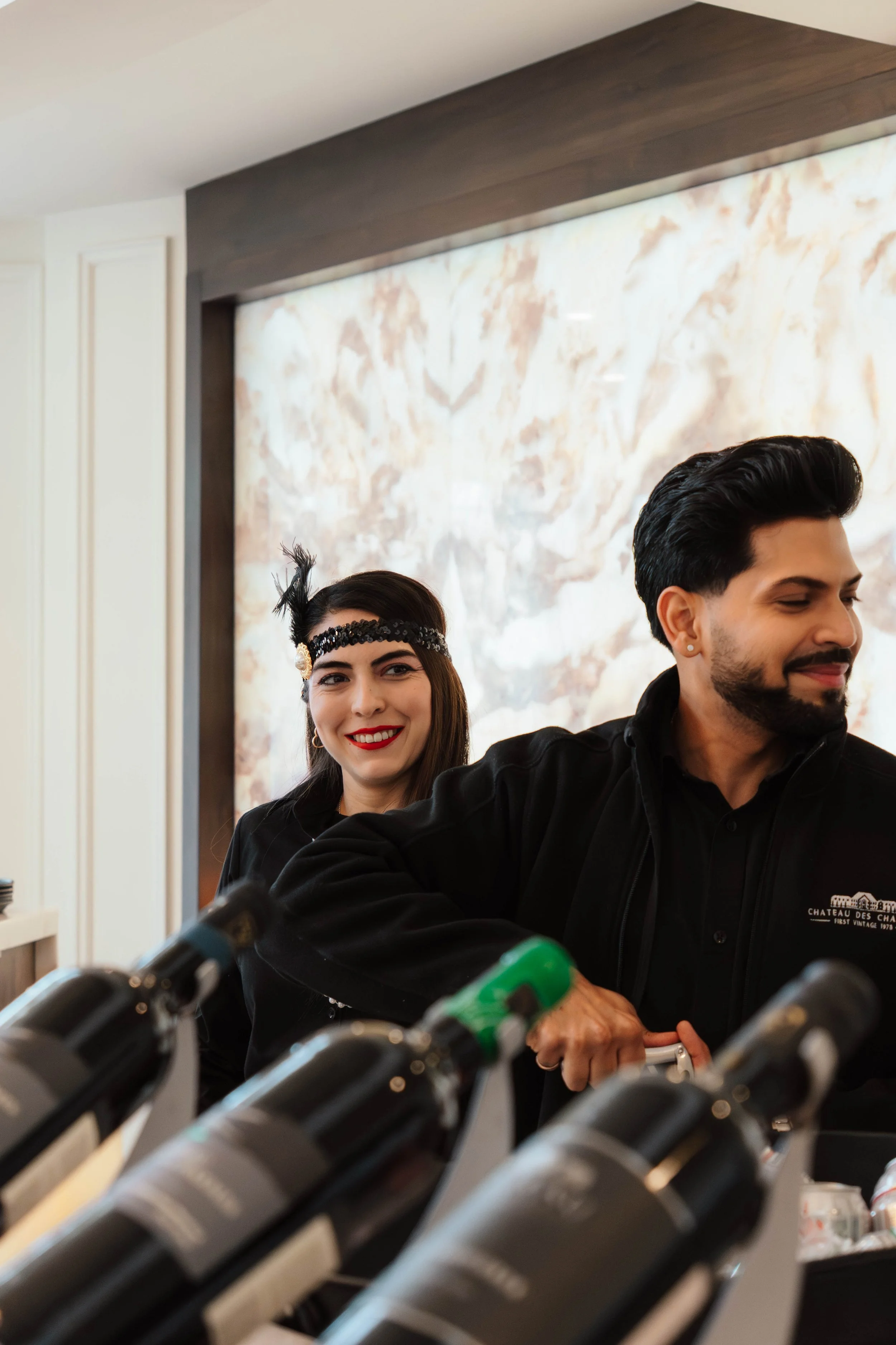 Two people pouring wine from bottles in a restaurant or bar setting. The woman is smiling with red lipstick, wearing a black outfit and a decorative headband. The man has dark hair and a beard, wearing a black jacket.