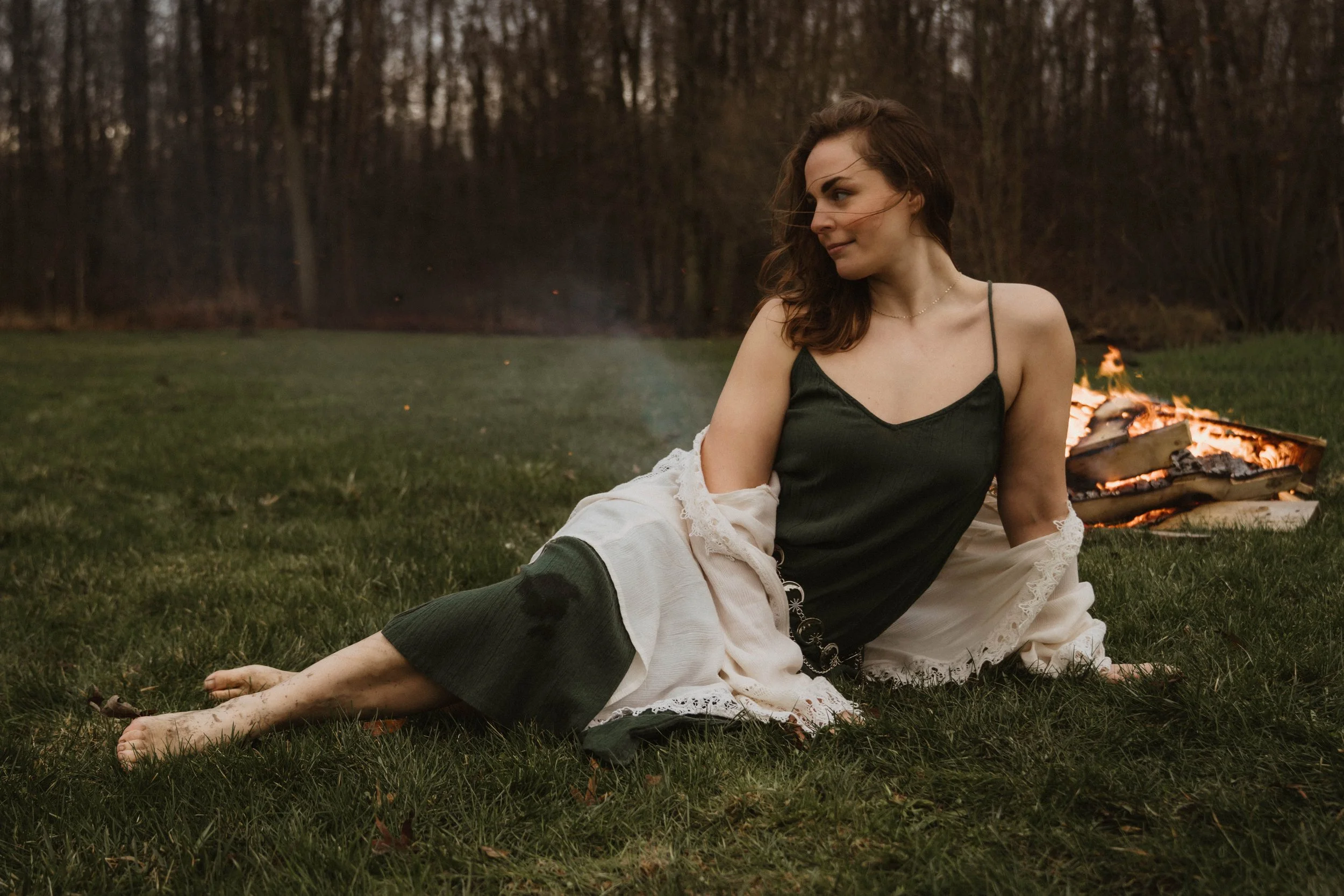 A woman in a dark green dress and white shawl sitting on grass near a small fire in a wooded area at dusk.