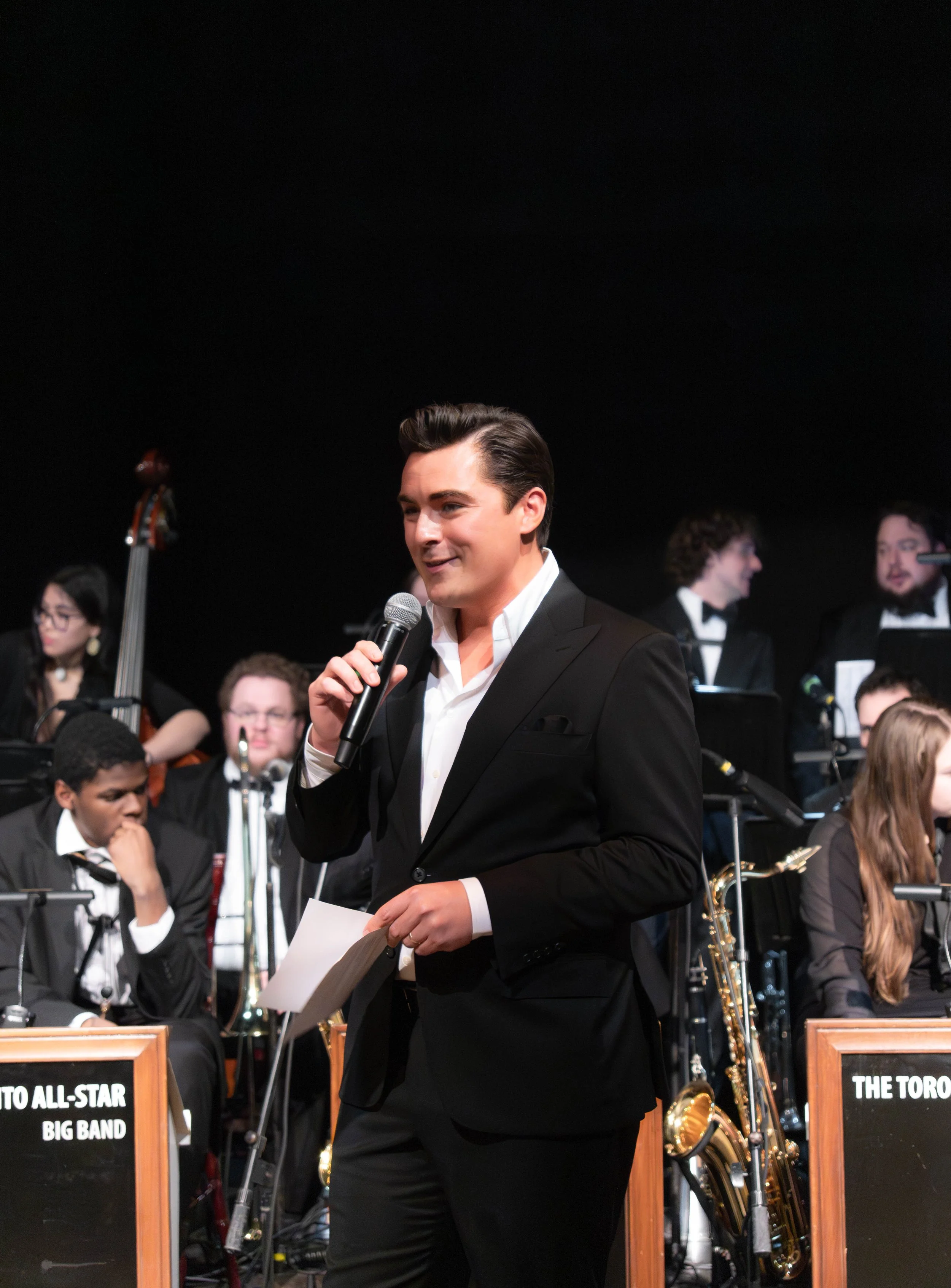 A man in a black tuxedo holding a microphone and speech notes, standing in front of an orchestra on stage.