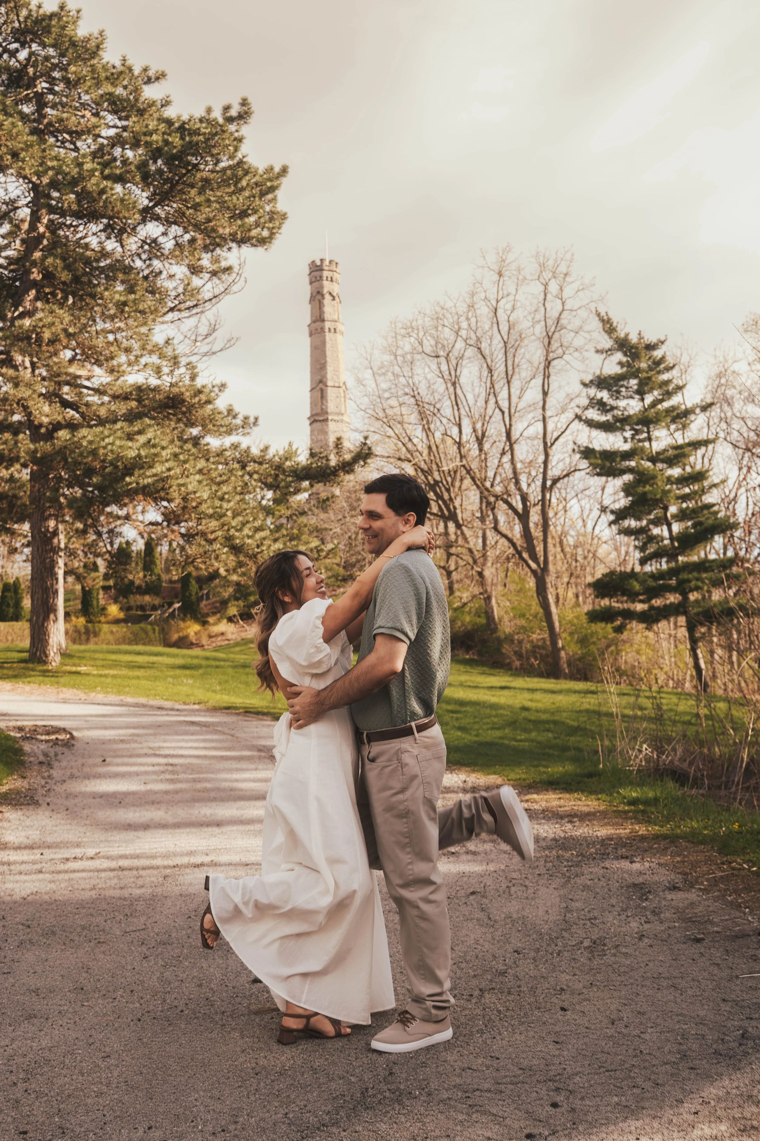A couple shares a joyful moment outdoors with trees and a tall tower in the background, one person kneeling while the other stands, both smiling.