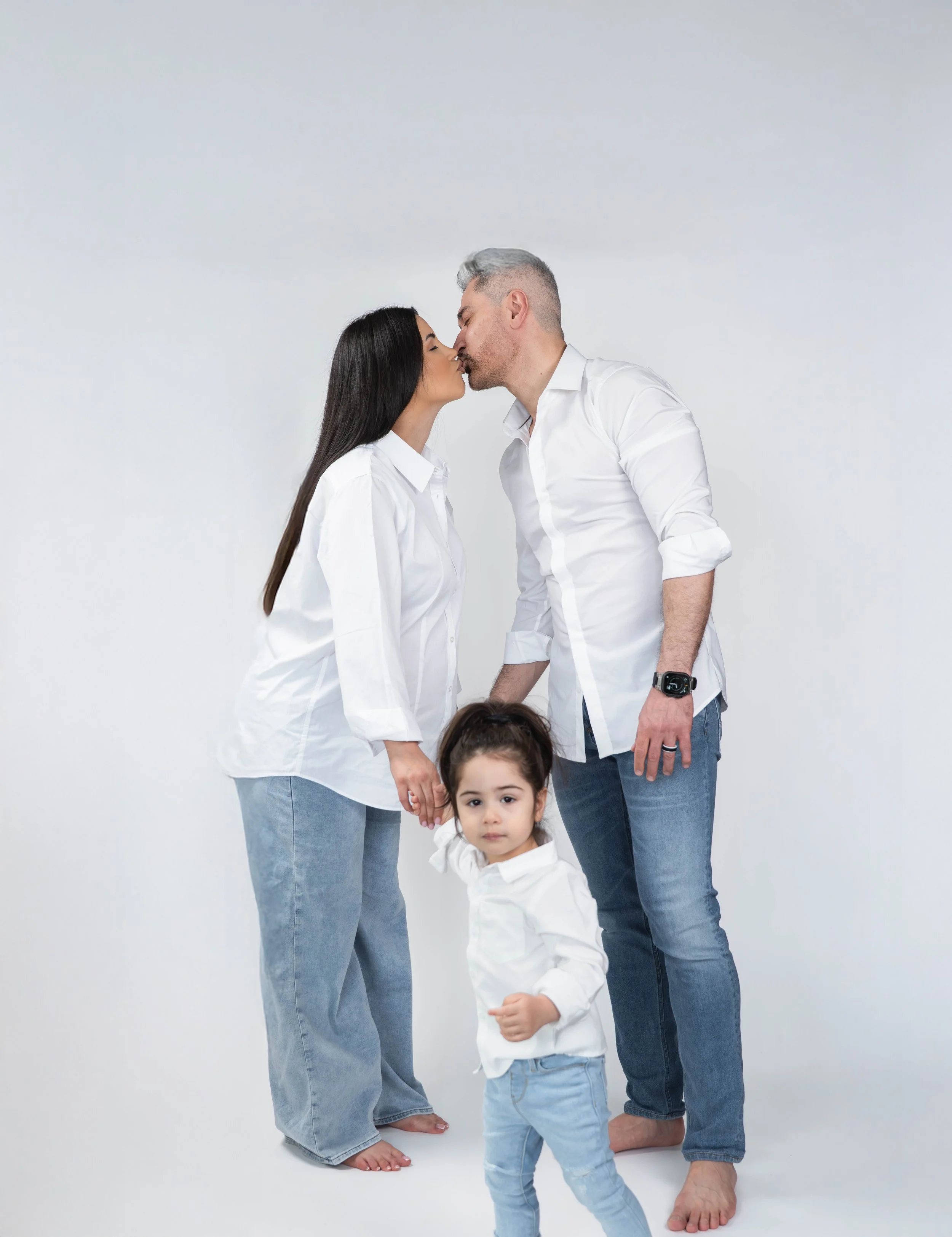 A family of four, including a man, woman, and two young children, are standing barefoot in front of a plain white background. They are holding hands, with the man and woman leaning toward each other for a kiss.