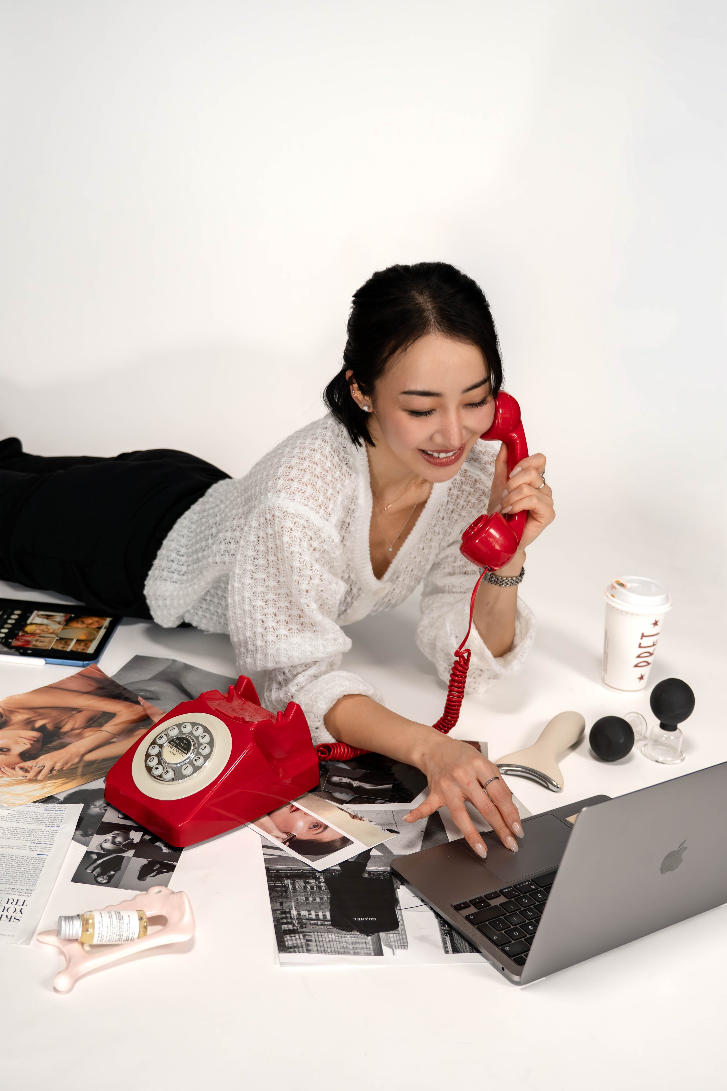 A woman lying on her stomach talking on a red rotary phone, surrounded by a laptop, magazines, cosmetics, and fitness equipment on a white table against a plain white background.