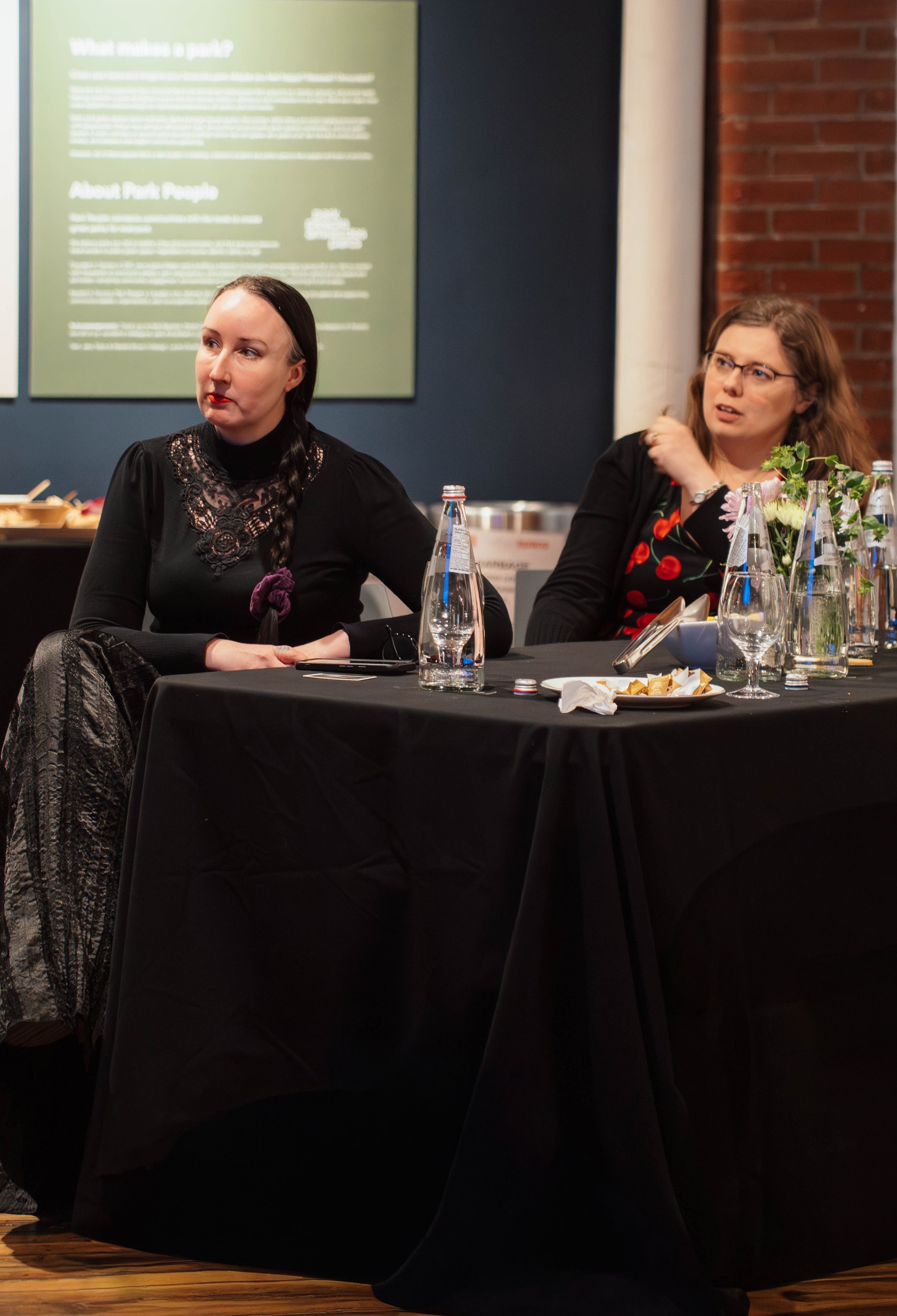Two women sitting at a black-covered conference table, engaged in discussion, with water bottles, glasses, a floral centerpiece, and a plate of snacks in front of them, against a background of a dark wall with informational posters.