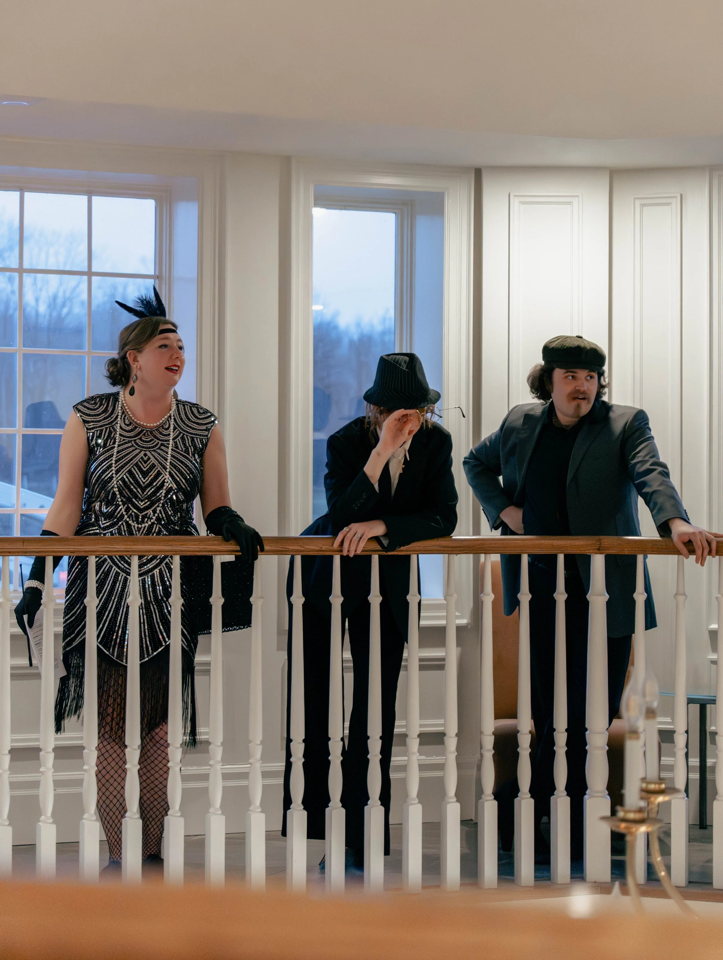 Three people dressed in vintage clothing standing on a balcony inside a house with large windows showing a wintery landscape outside.