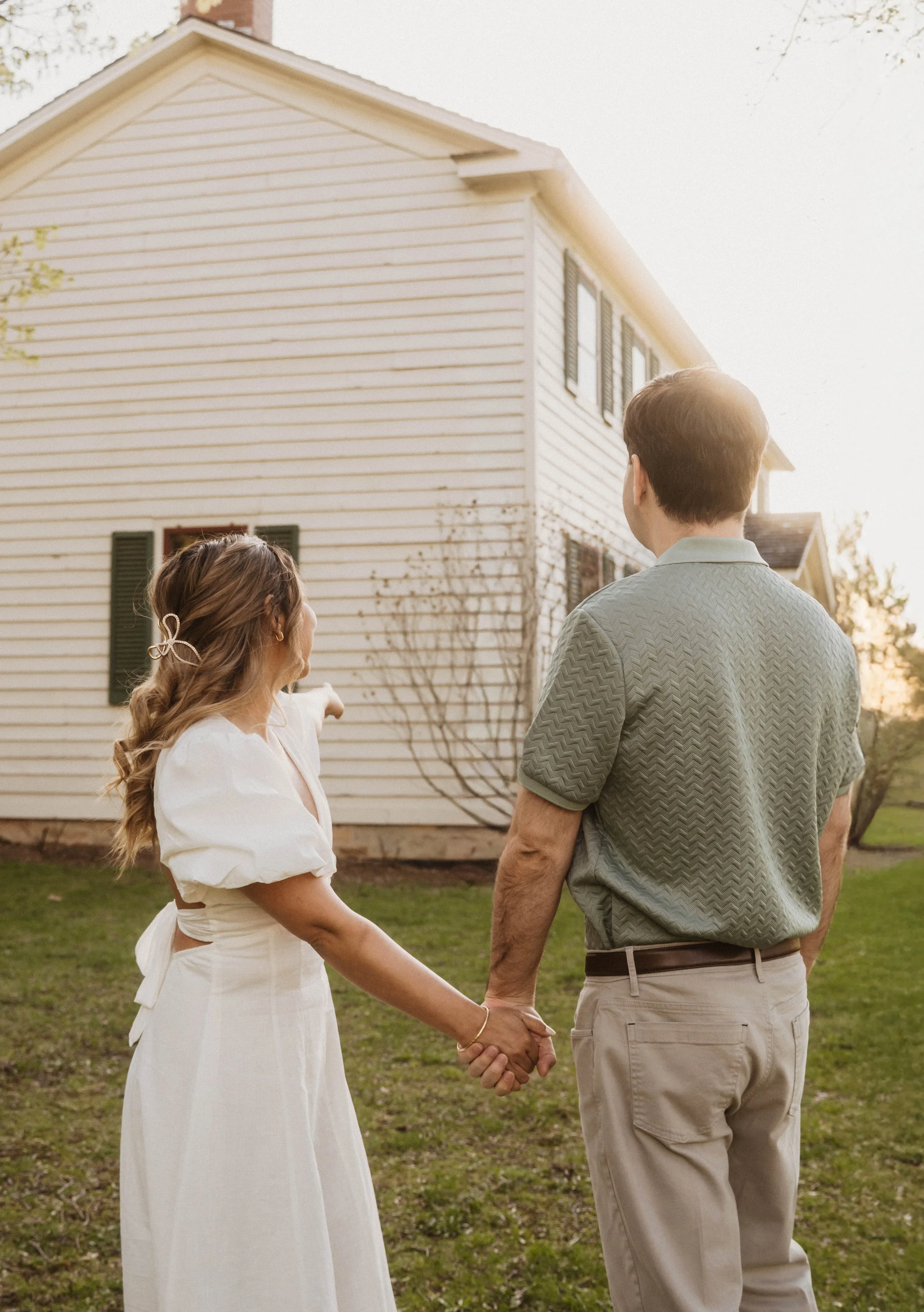 A couple standing outdoors holding hands during sunset, the woman wearing a white dress with puffed sleeves and the man in a patterned green shirt and beige pants, in front of a white house with green shutters.