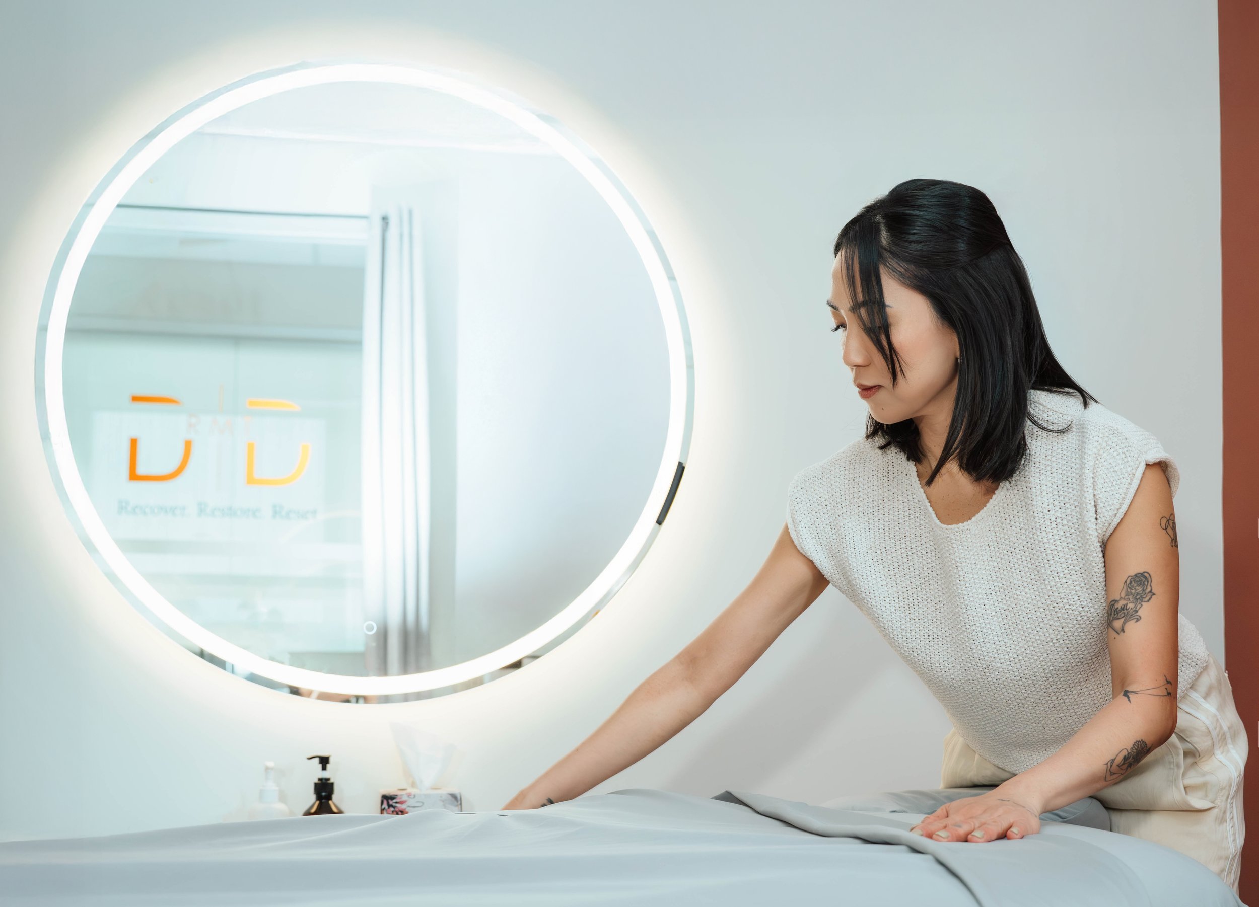 Woman with black hair, wearing a white sleeveless top, arranging a gray sheet on a table in front of a backlit round mirror in a modern, minimalistic room.