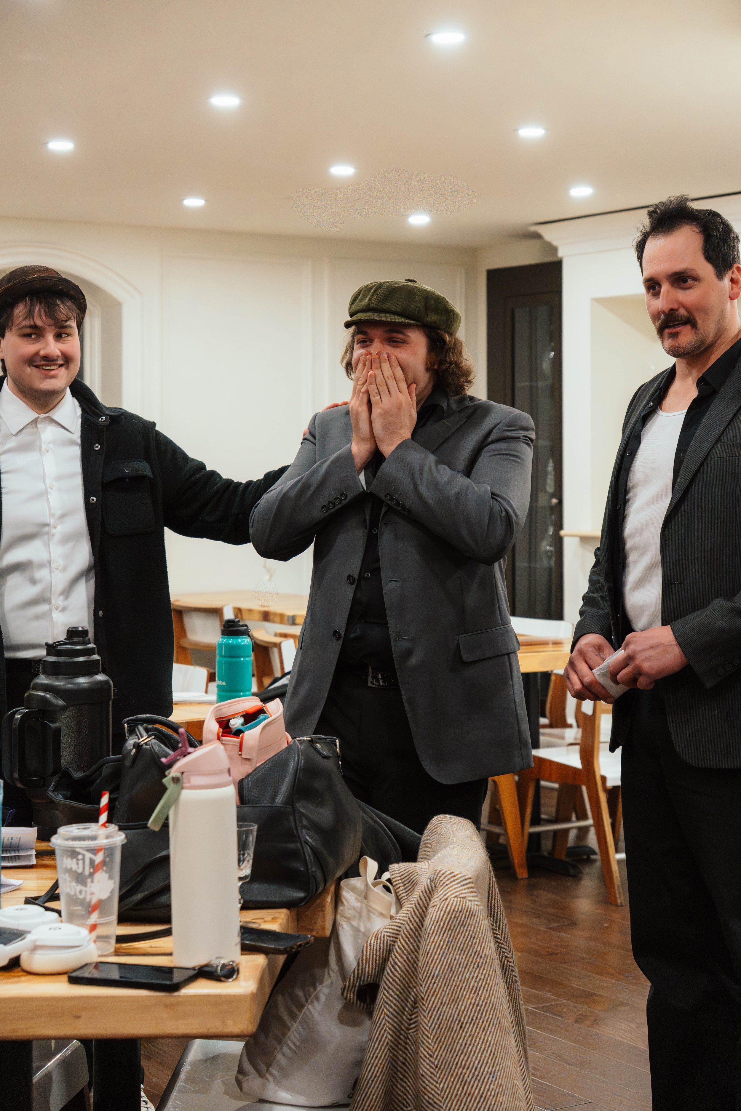 Three men standing around a table, one with an arm on another's shoulder, the middle man expressing surprise or excitement with hands covering mouth, in a cozy dining room.