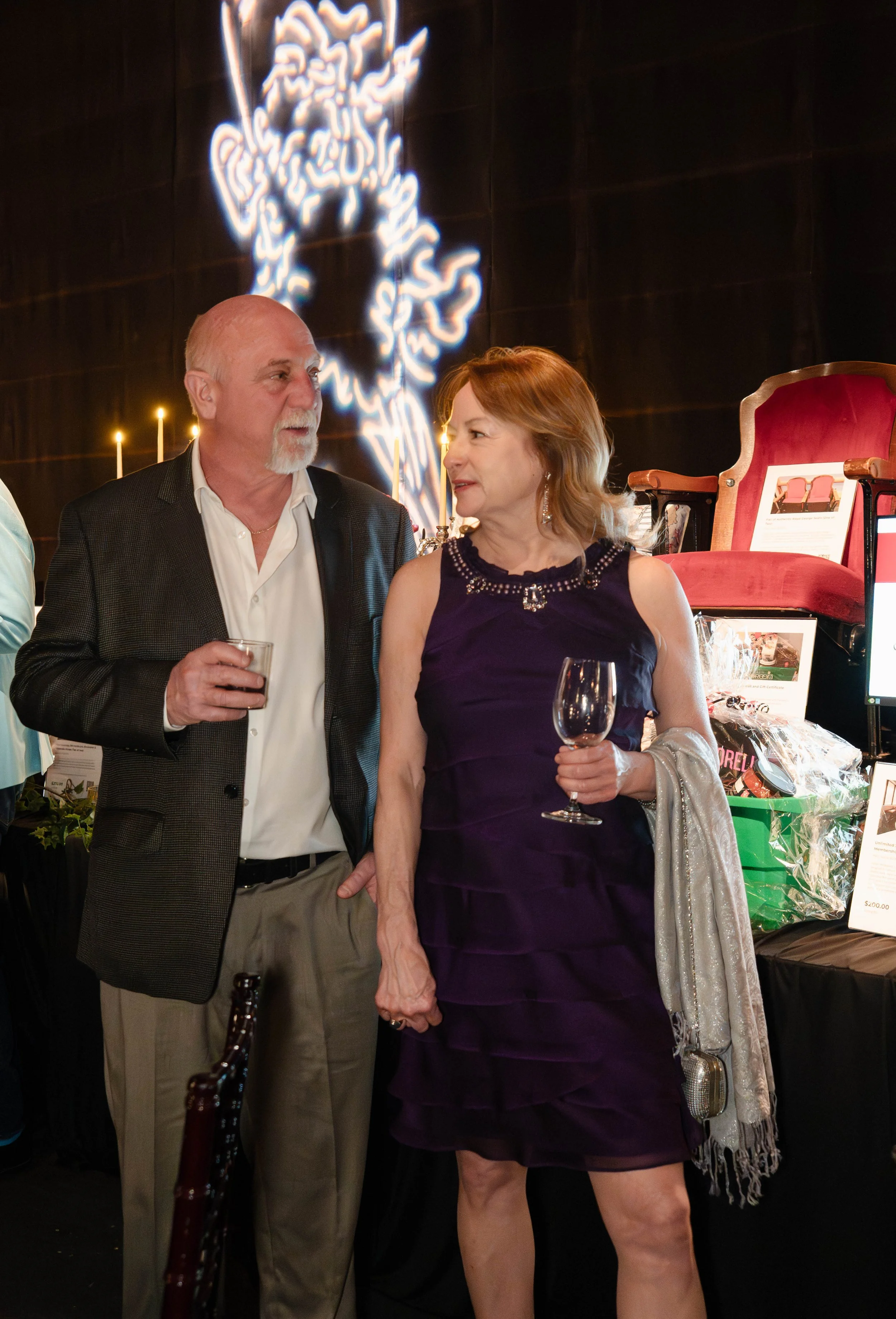 A man and a woman are engaged in conversation at an indoor event. The man has a bald head, a beard, and is wearing a dark blazer over a white shirt. The woman has red hair, and is wearing a dark purple dress with decorative details on the neckline, h