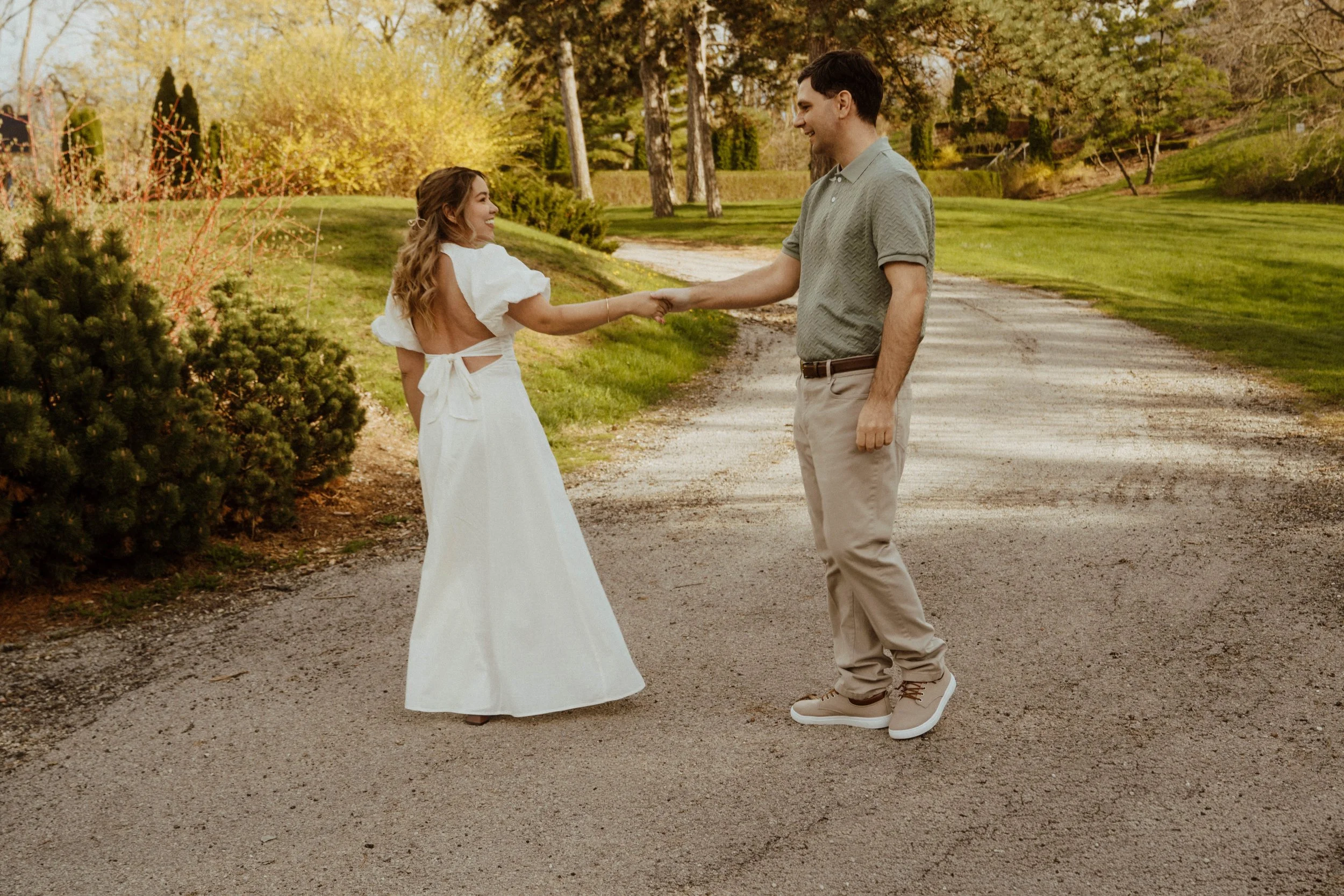 A couple holding hands and smiling outdoors on a dirt path, with trees and greenery in the background during late afternoon or early evening.