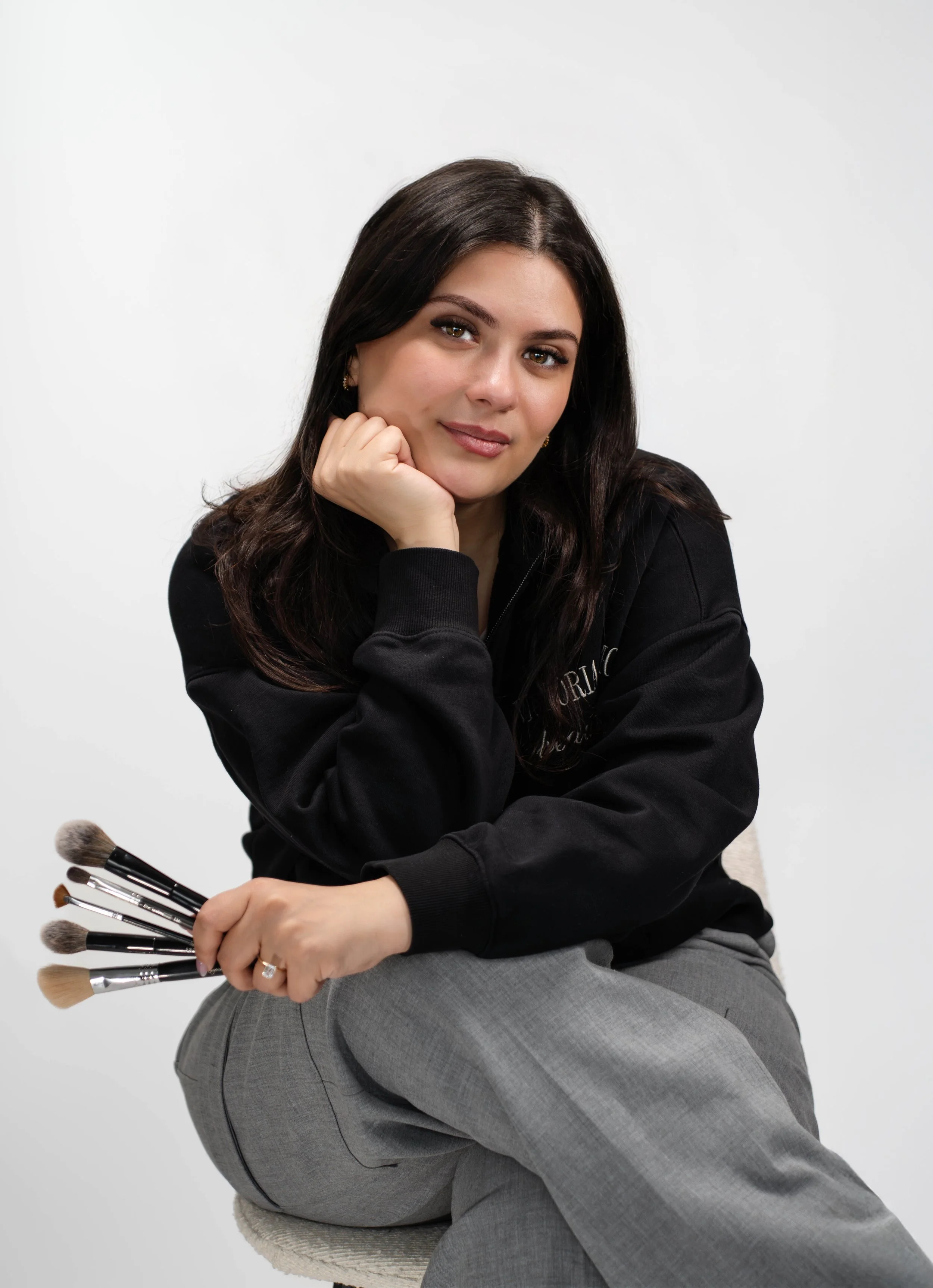 A woman with dark brown hair, wearing a black sweatshirt and gray pants, sitting on a chair. She is resting her chin on her left hand, and holding a bunch of makeup brushes in her right hand.