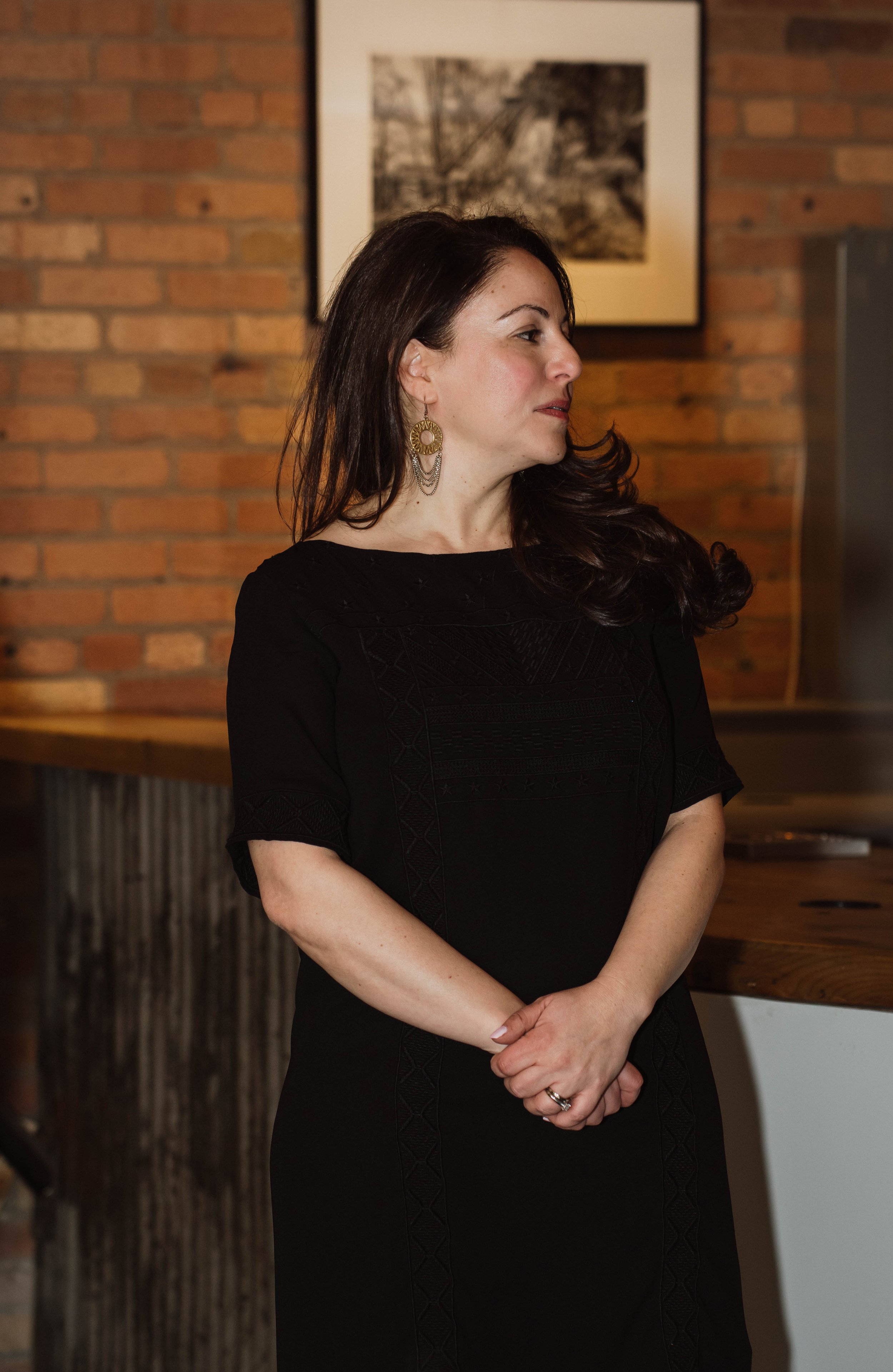 A woman with dark brown hair and earrings wearing a black dress, standing indoors against a brick wall background with framed pictures.