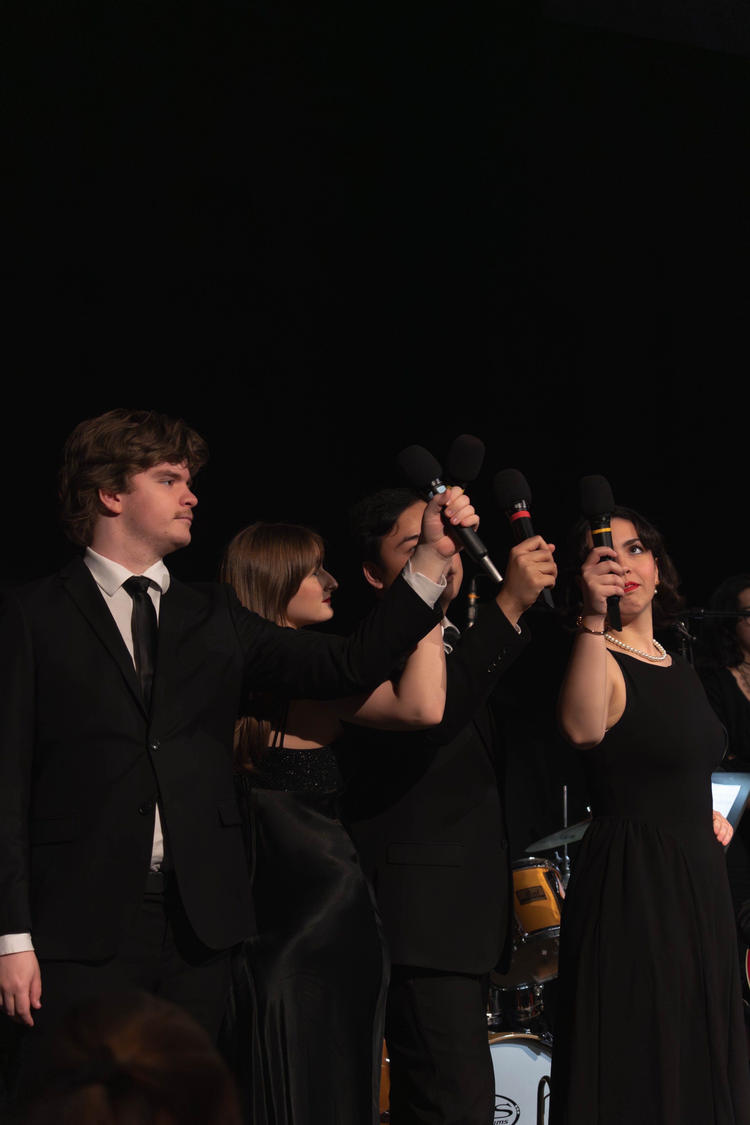 Four formally dressed people, holding microphones, standing on a stage against a black background.
