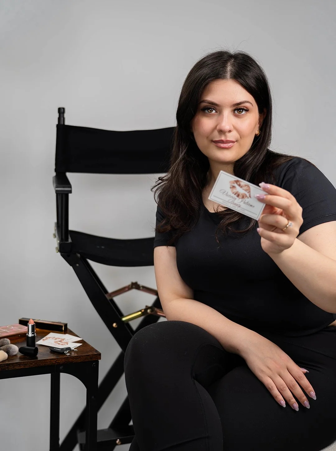 A woman with dark hair holding a business card, sitting beside makeup products on a table, with a director's chair in the background.