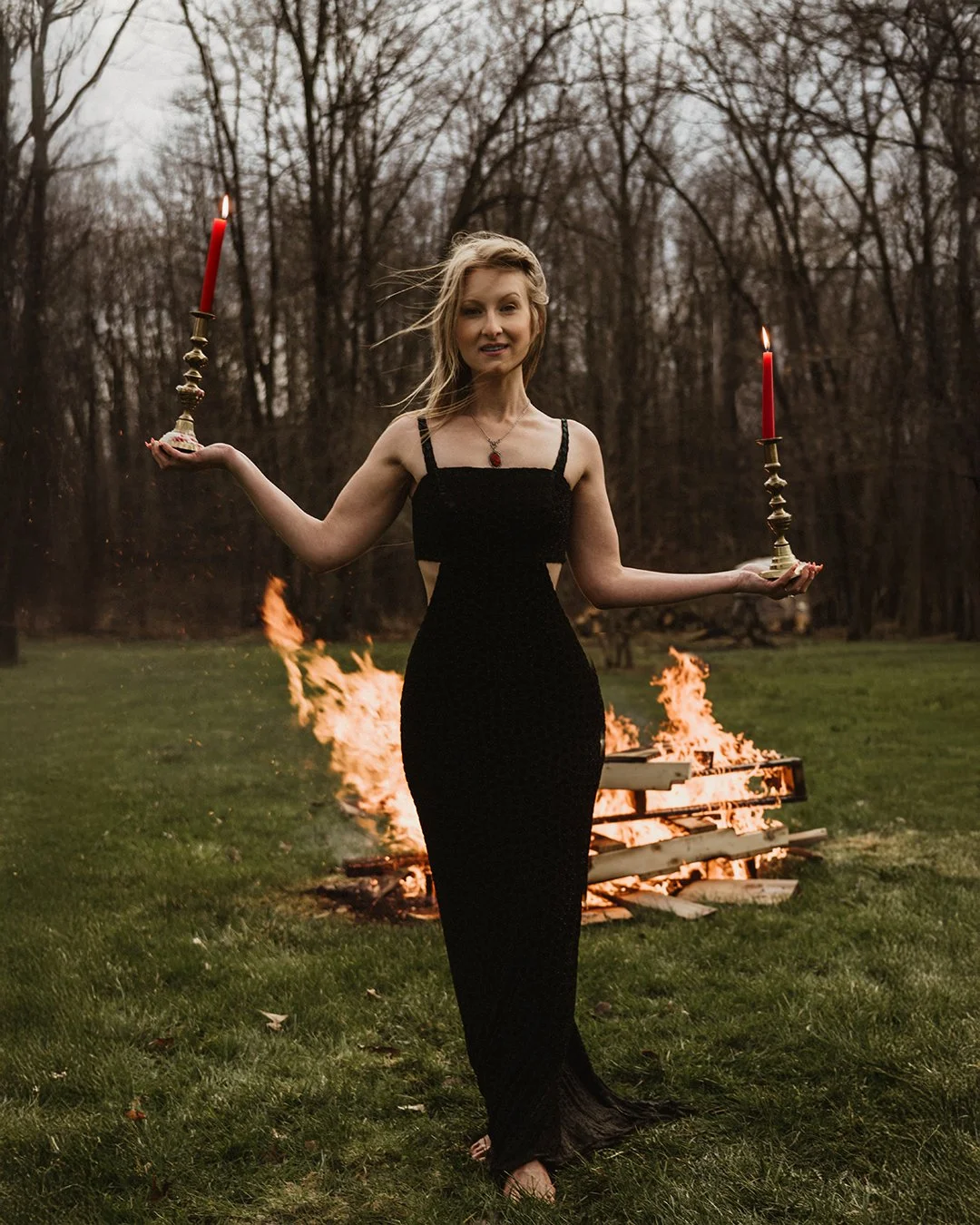 A woman in a black dress standing outdoors in front of a burning bonfire, holding two candlesticks with lit red candles, one in each hand.