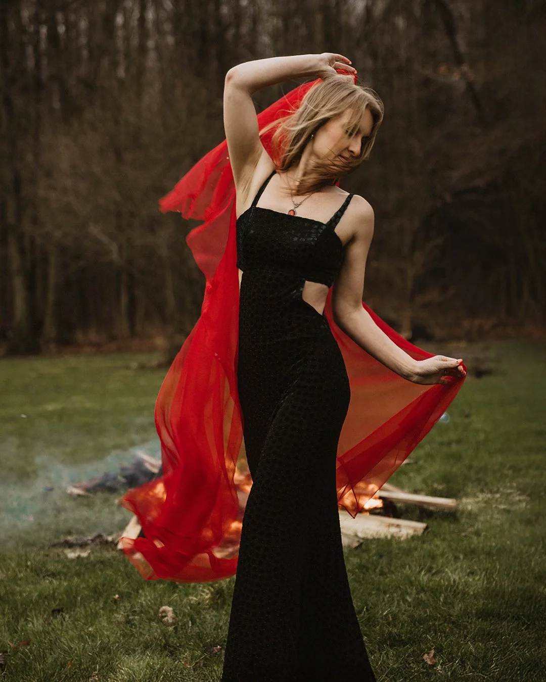 A woman in a black dress standing outdoors with a red sheer fabric flowing around her, holding it with both hands, with trees in the background.
