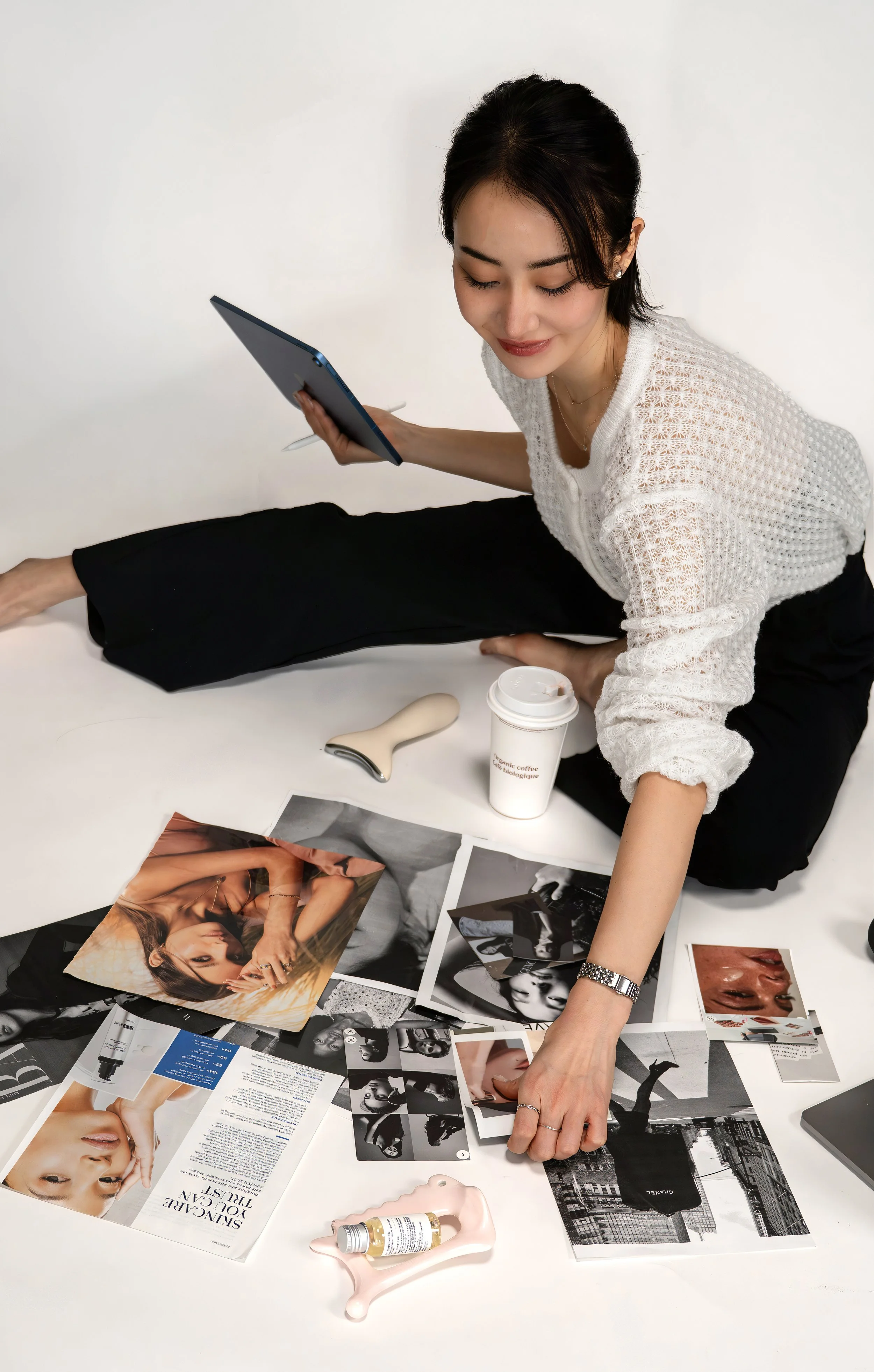 A woman sitting on the floor surrounded by fashion photographs, skincare products, and a coffee cup, holding a tablet.