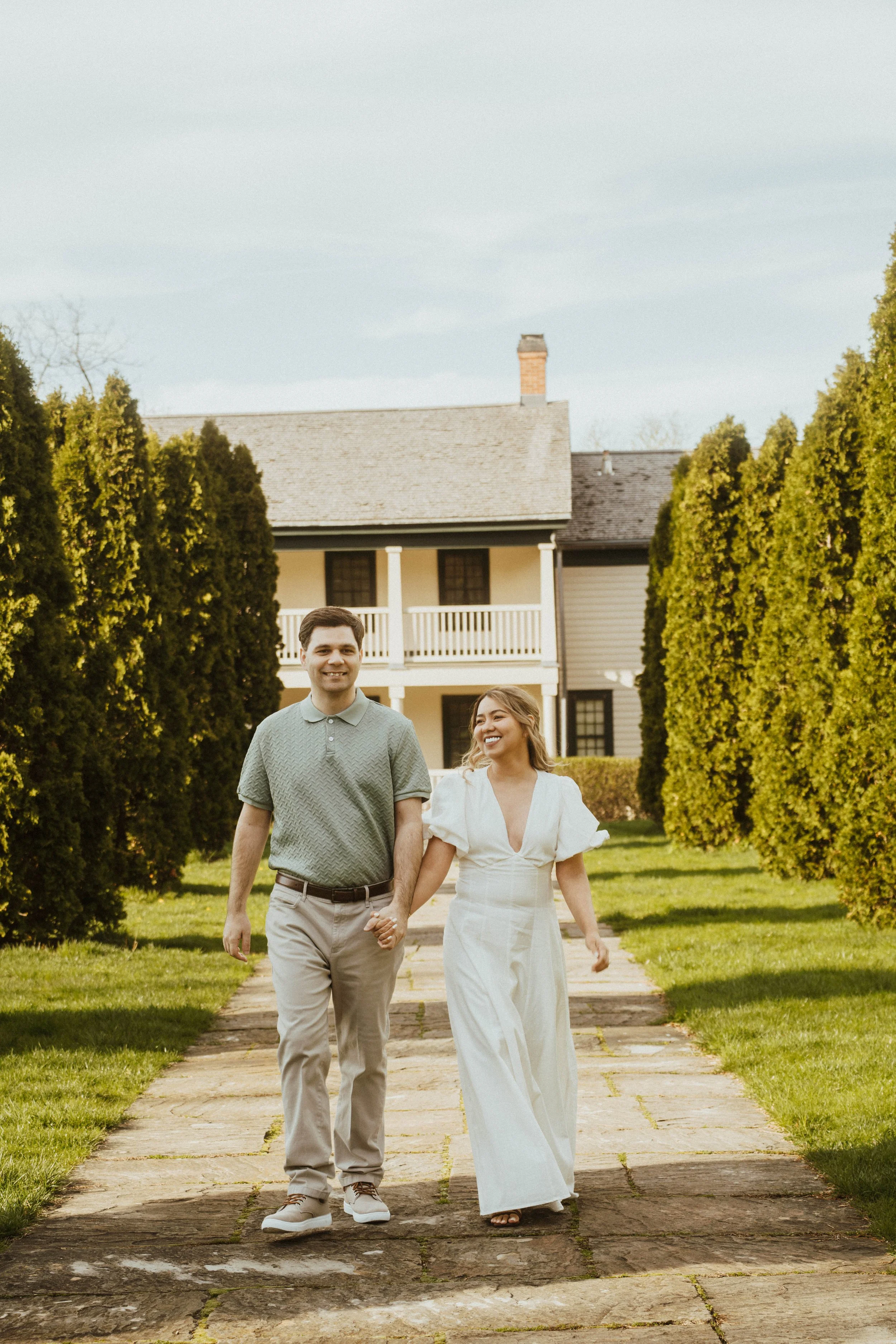 A smiling man and woman holding hands and walking down a stone-paved path surrounded by tall green hedges, with a white house and trees in the background on a sunny day.